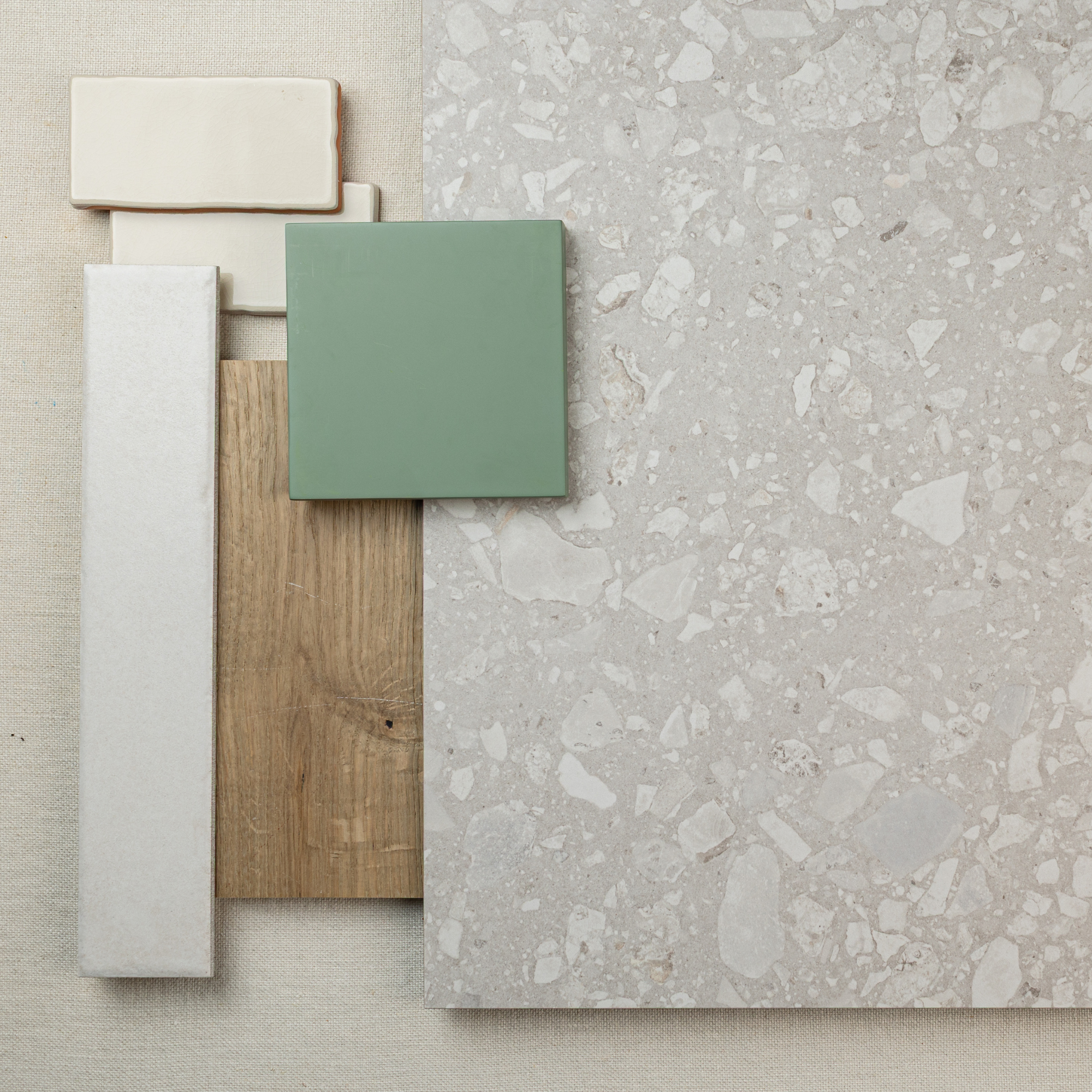 Flat lay of soft green ceramic tile, natural wood plank, beige rectangular tiles, and large light terrazzo tile on fabric surface
