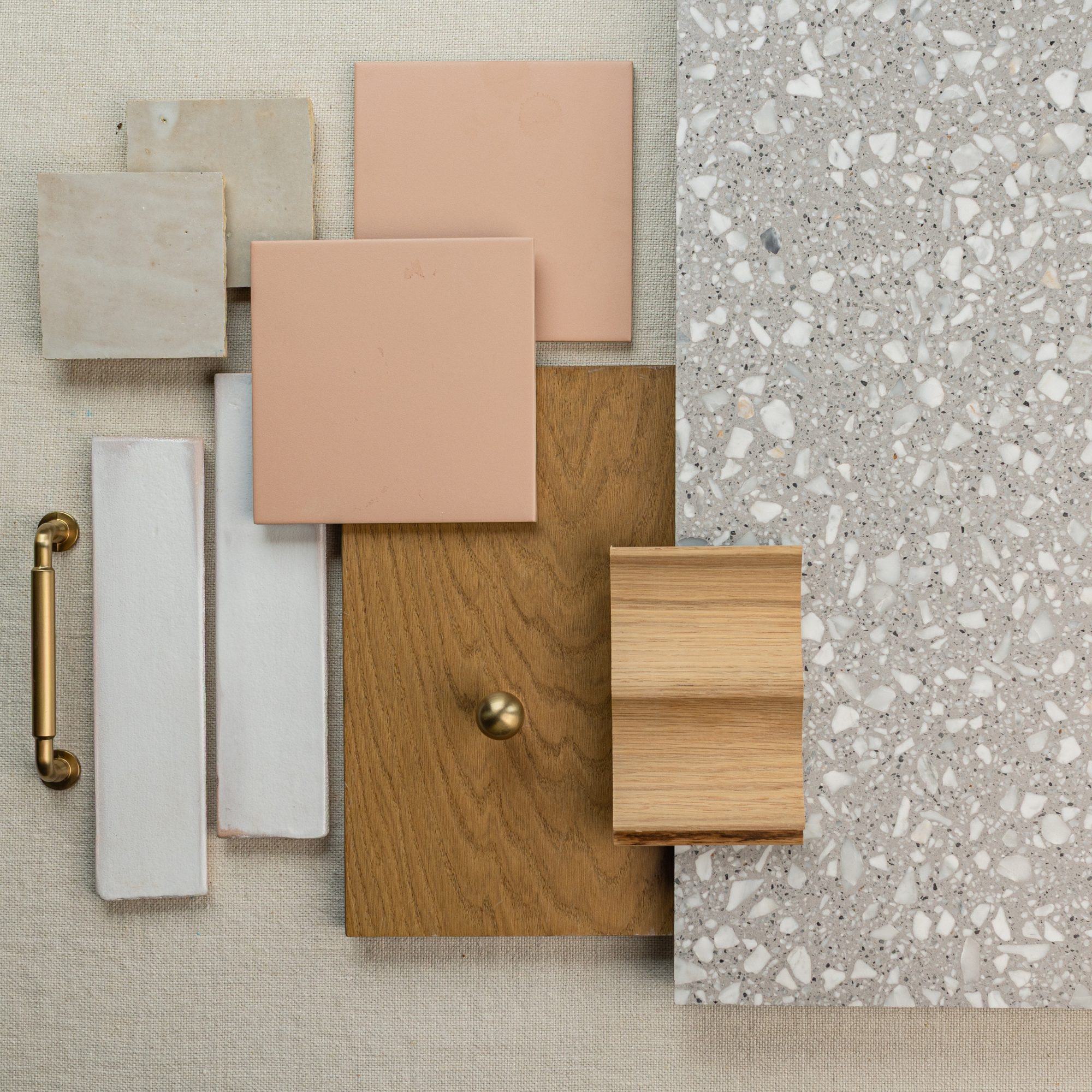 Selection of matte pink square tiles, natural wood samples, terrazzo surface, elongated white ceramic tiles, and brass hardware on beige fabric background