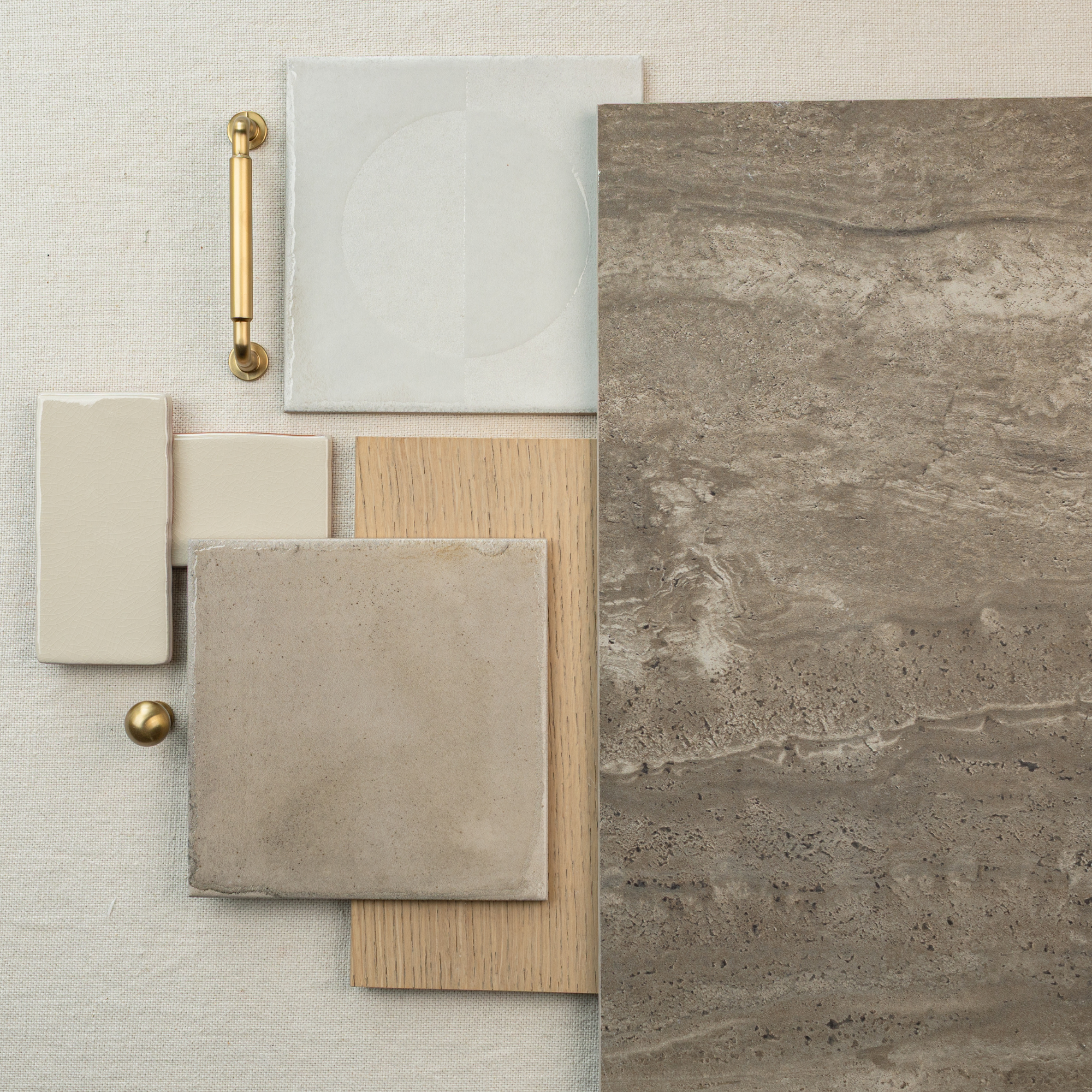 Flat lay of neutral stone-look large format tile, beige ceramic tiles, light wood plank, and brass hardware on textured fabric background