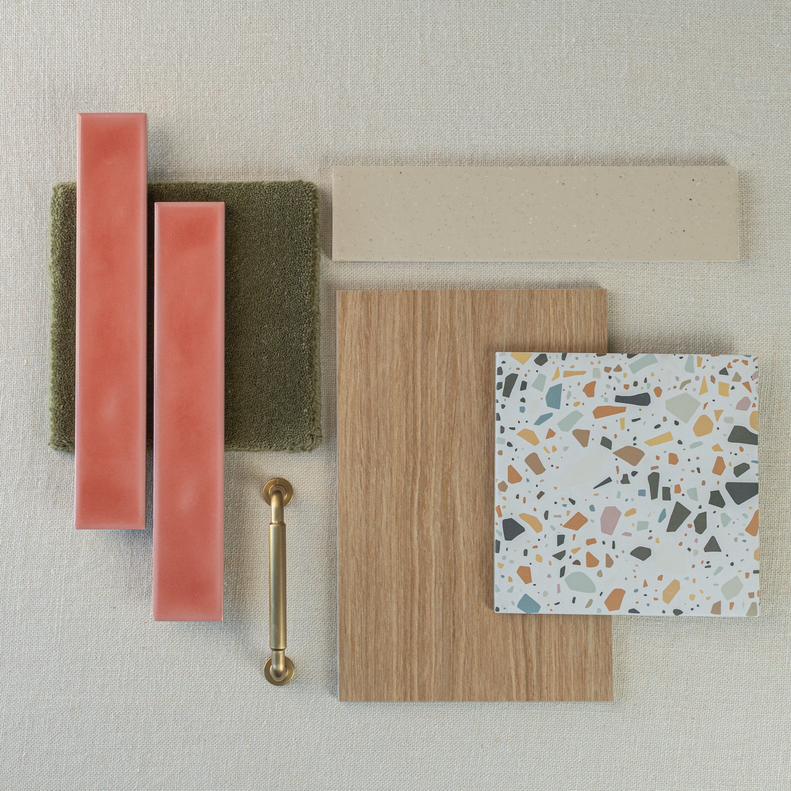 Flat lay of pink ceramic tiles, green textured tile, oak wood sample, terrazzo tile, cream tile strip, and brass cabinet pull on fabric