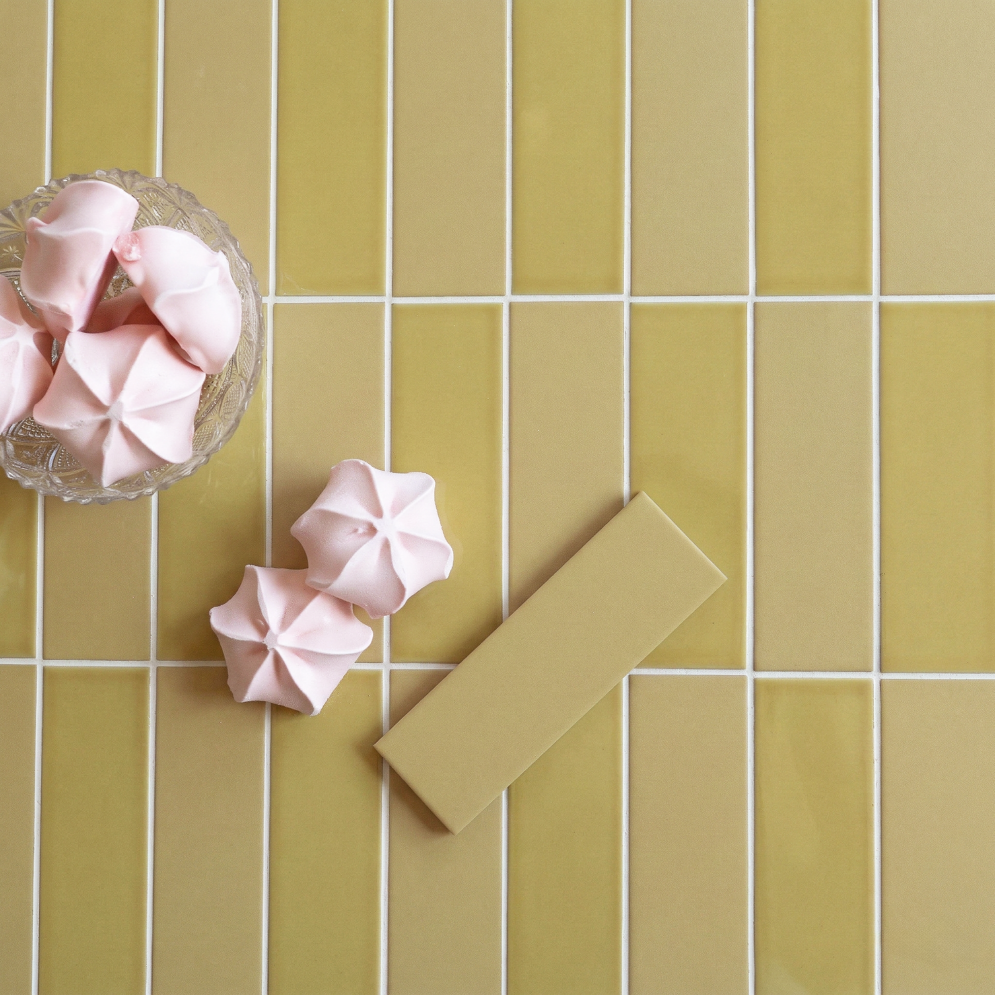 Yellow rectangular tiles with white grout, a matching yellow tile sample, and pink meringue cookies on a glass dish and surface