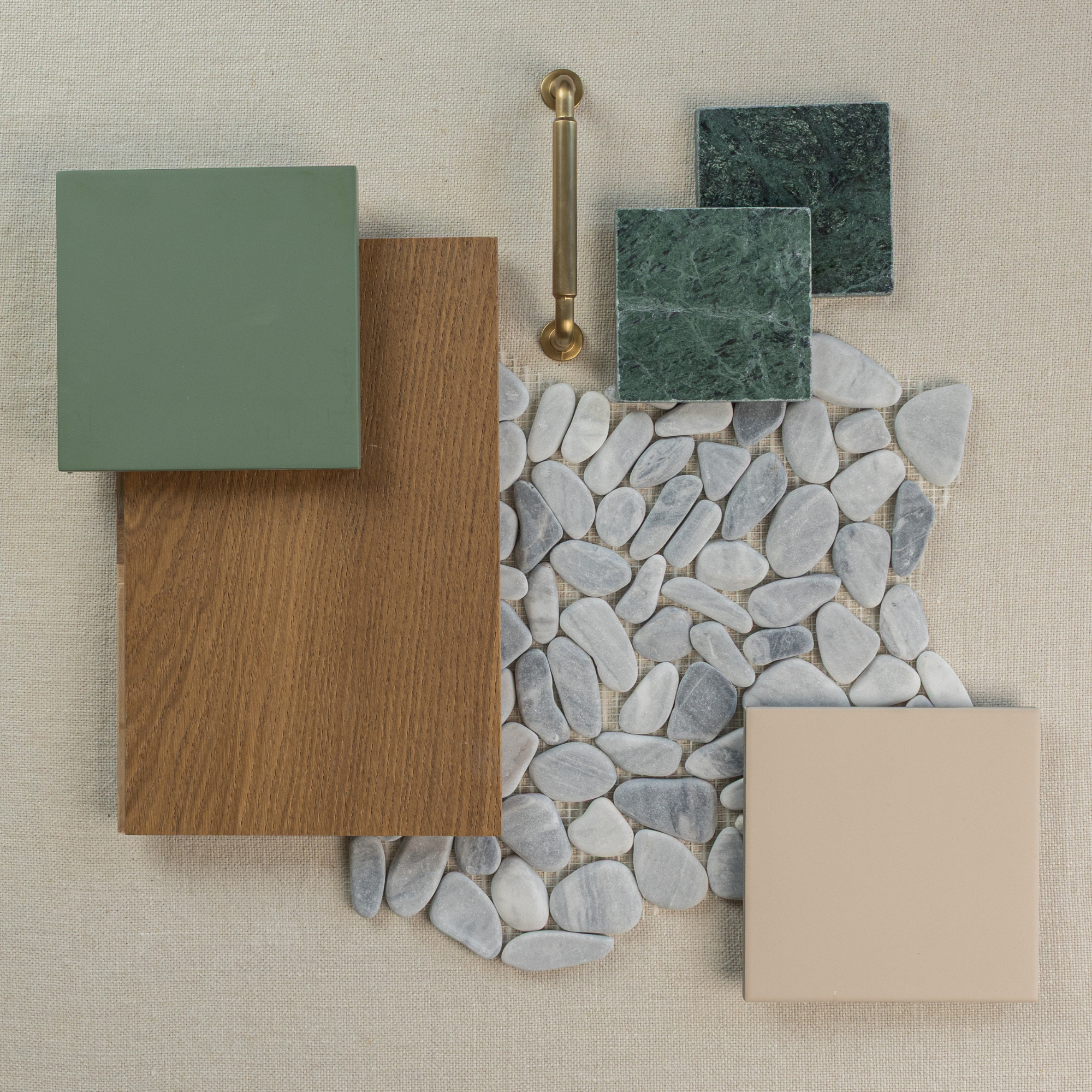 Flat lay of smooth white pebbles surrounded by oak wood, green and beige ceramic tiles, green stone samples, and brass handle on fabric background