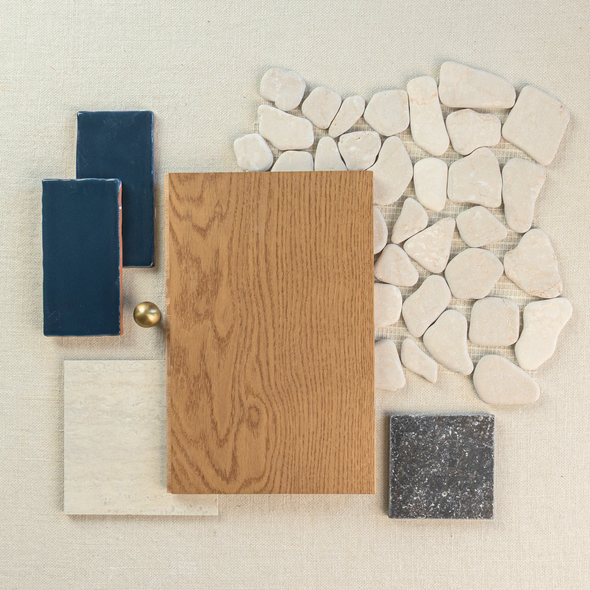 Flat lay of natural white stone pebbles with oak wood sample, deep blue ceramic tiles, textured gray tile, and cream ceramic swatch on beige fabric