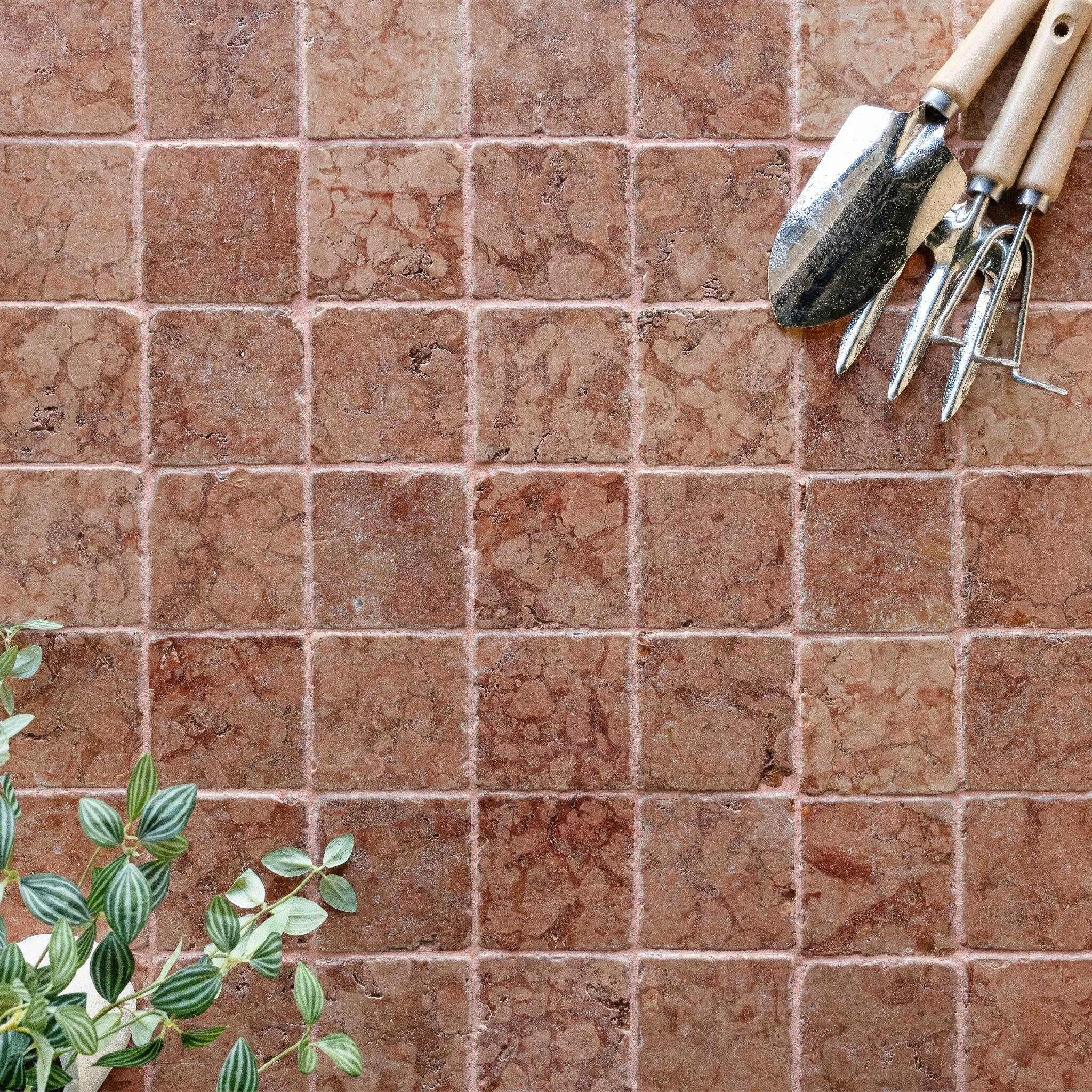 Square tumbled stone tiles in warm reddish-brown tones with gardening tools and green leafy plant nearby