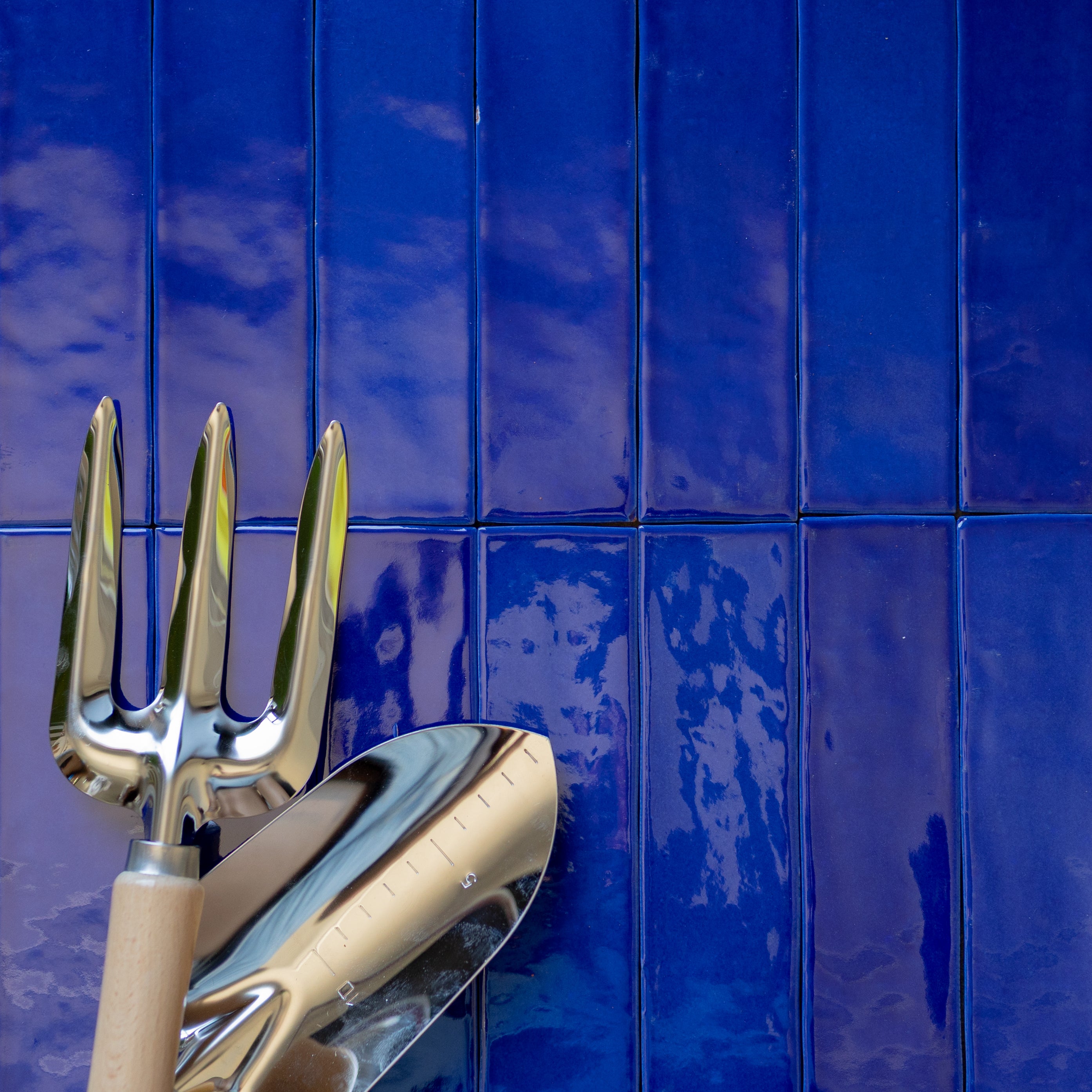 Glossy sapphire blue ceramic tiles with vertical rectangular shape and reflective gardening tools with wooden handles resting on them