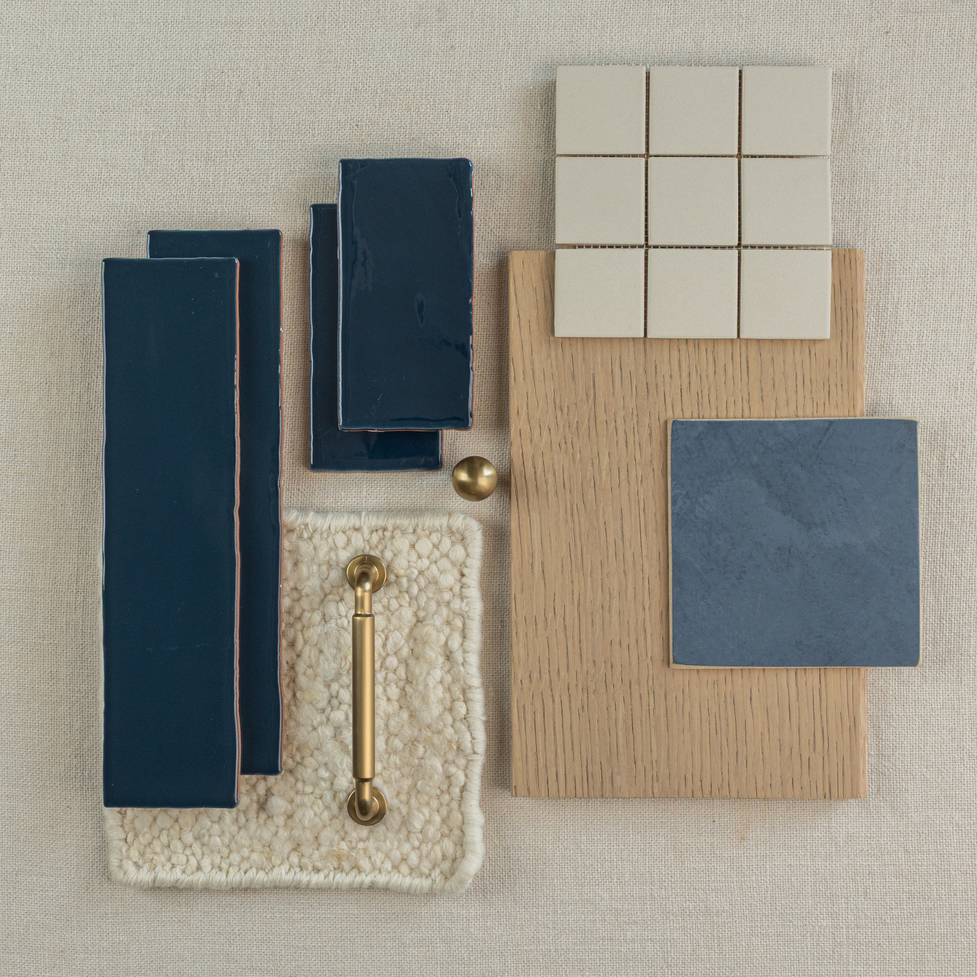 Flat lay of glossy deep navy rectangular tiles, matte beige square tiles, wood sample, textured cream fabric, brass handle, and dark blue ceramic tile