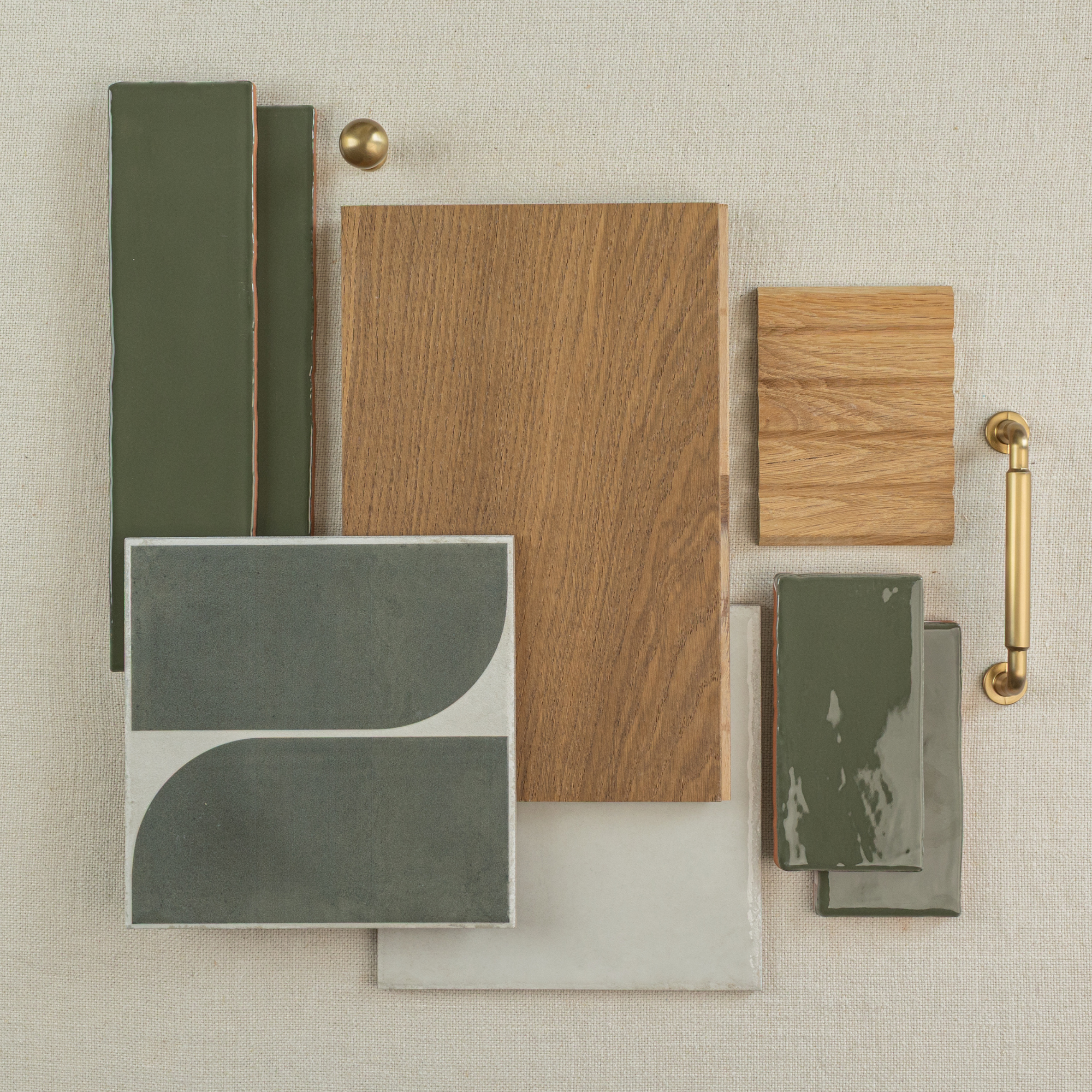 Flat lay of glossy olive green rectangular tiles, wood samples, matte gray and white tiles, and brass hardware on neutral fabric