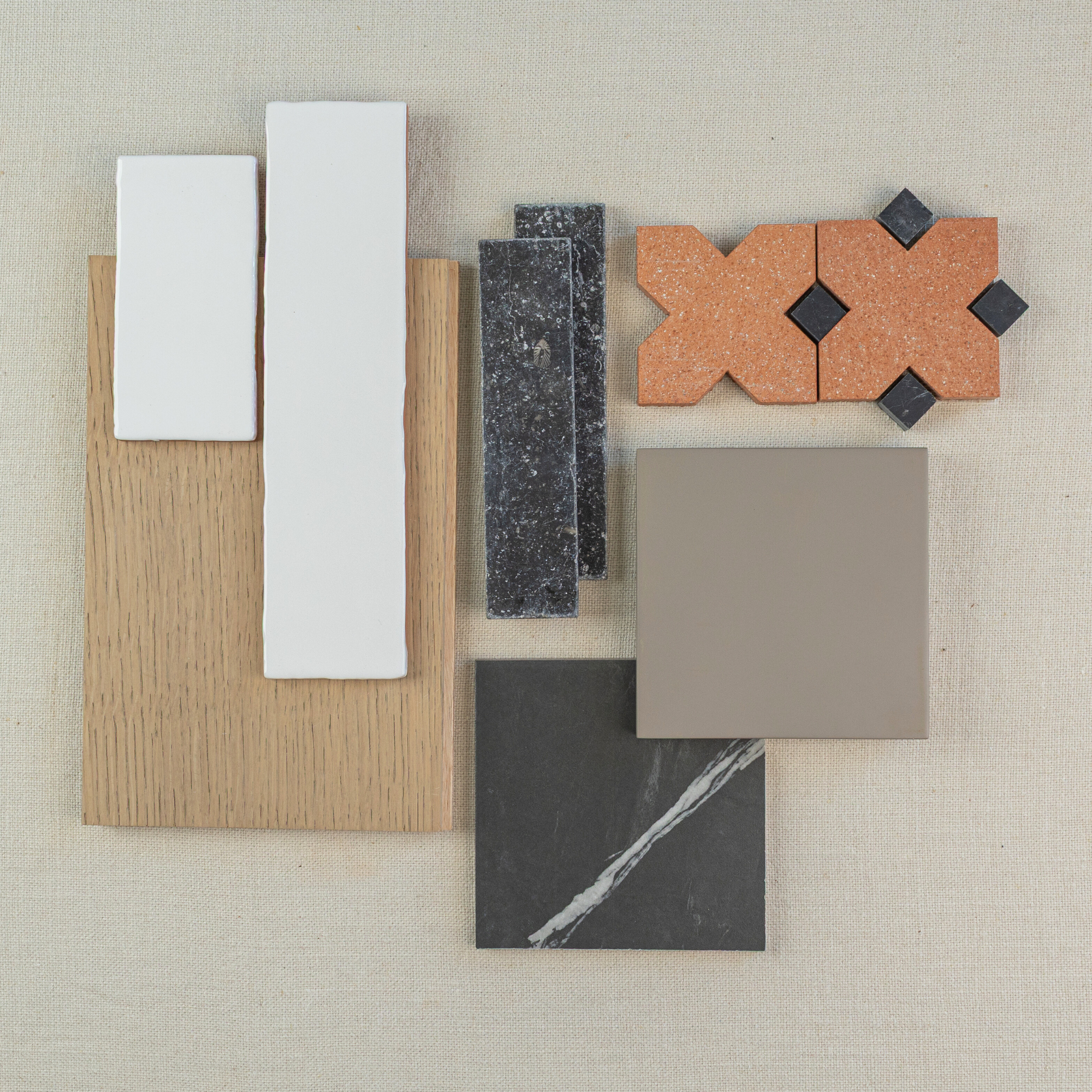 Flat lay of white ceramic tiles, terracotta patterned pieces, dark marble, taupe square tile, and oak wood sample on neutral fabric