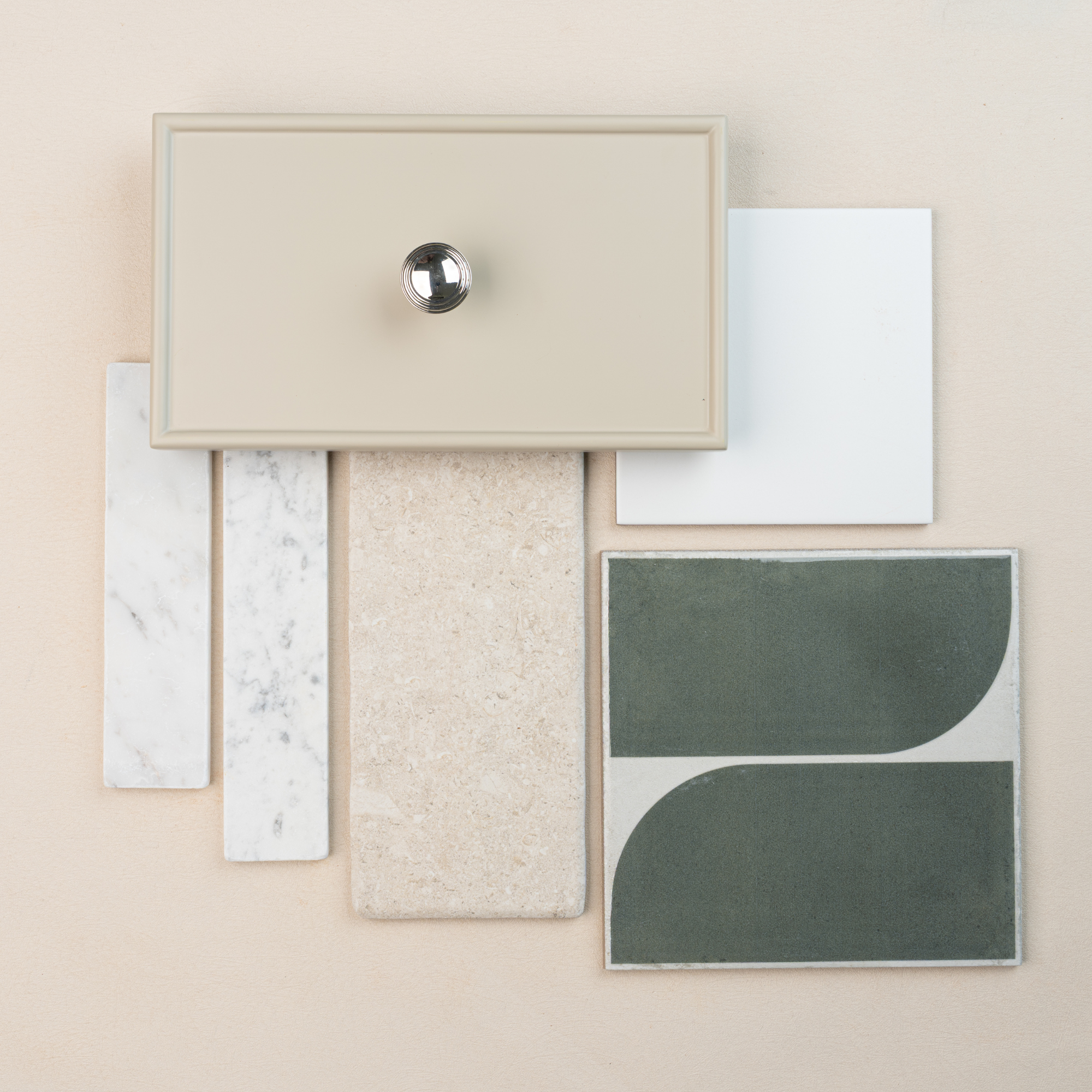 Flat lay of beige cabinet drawer front with chrome knob, green curved ceramic tile, white square tile, and stone tile samples on pale background
