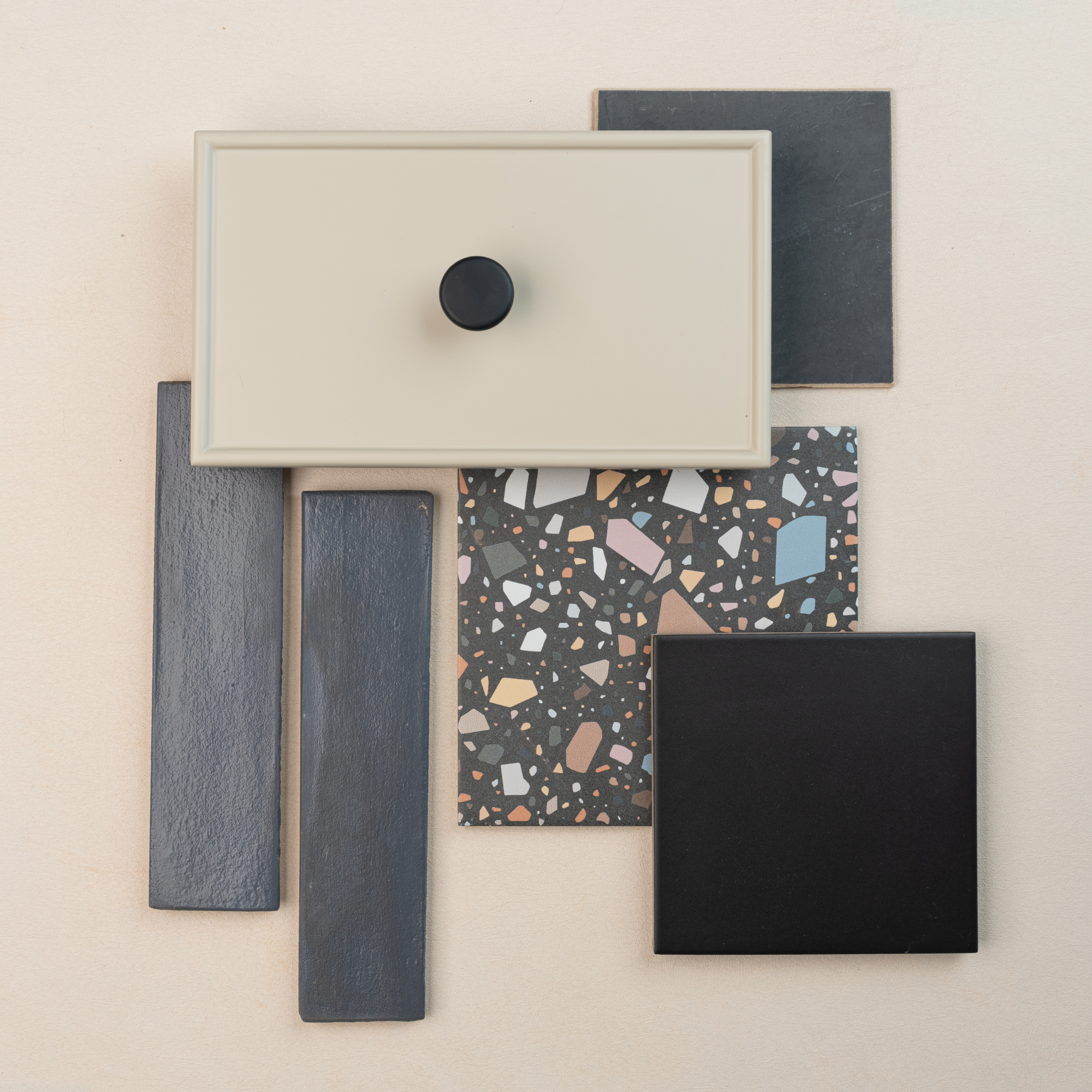 Minimalist beige cabinet drawer front with black knob atop terrazzo tile and dark gray and black ceramic tile samples on pale surface