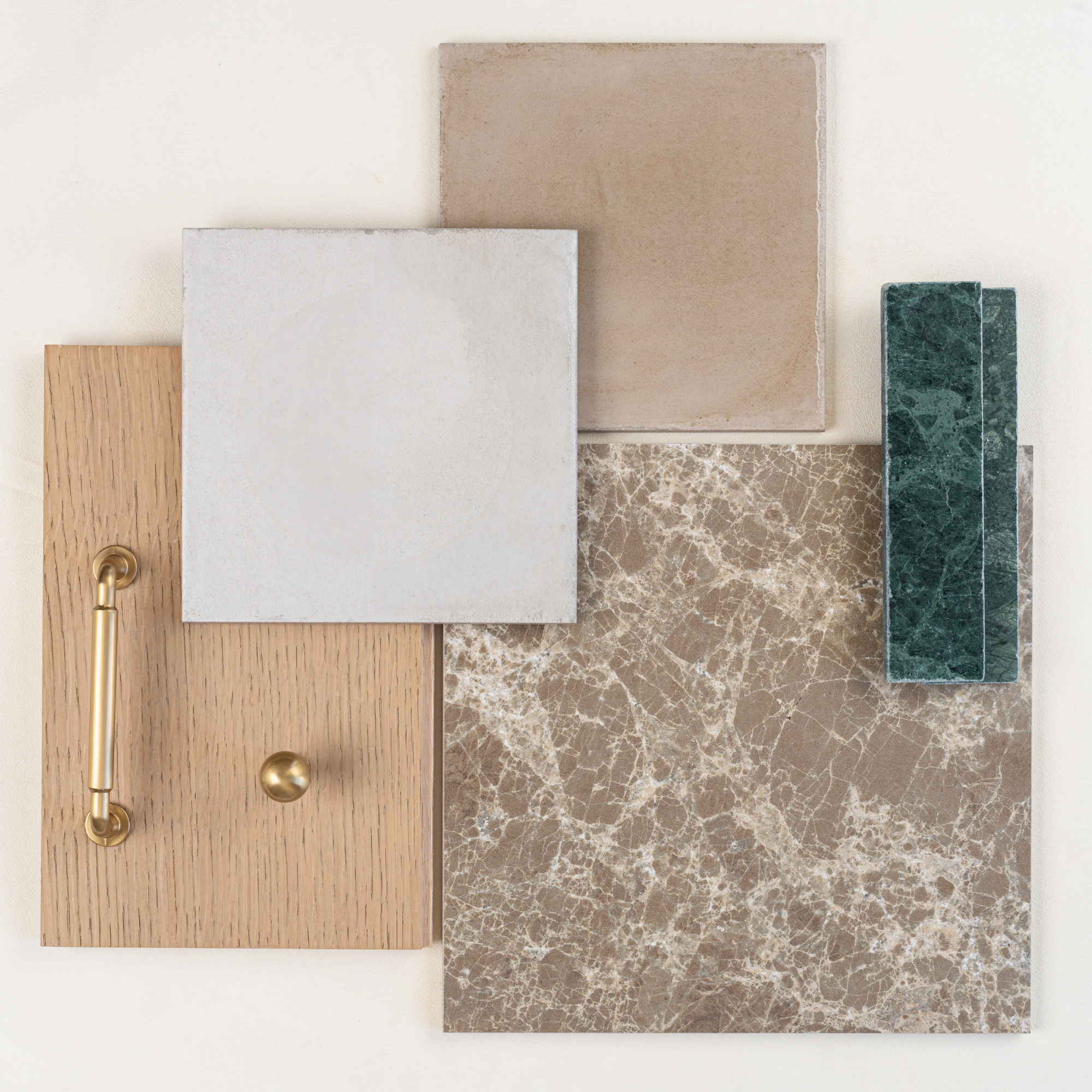 Top view of beige ceramic, brown marble, green stone tiles, and oak wood samples with brass handle and knob arranged on pale background