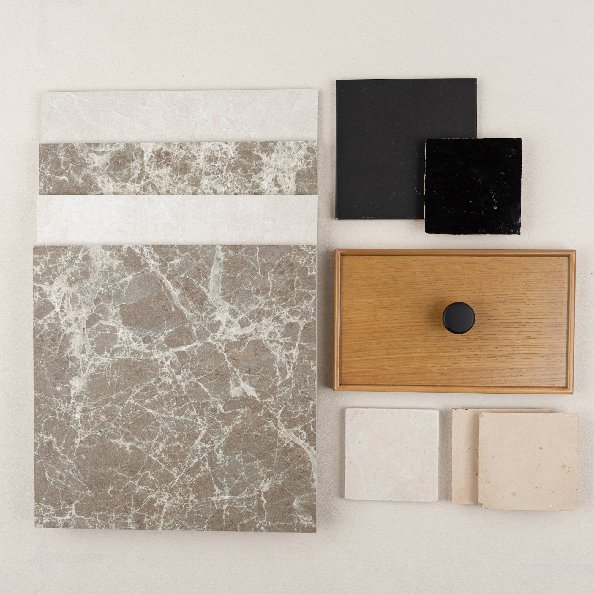 Arrangement of marble tile samples, black stone squares, light beige textured tiles, and a wood drawer front with black knob on pale background