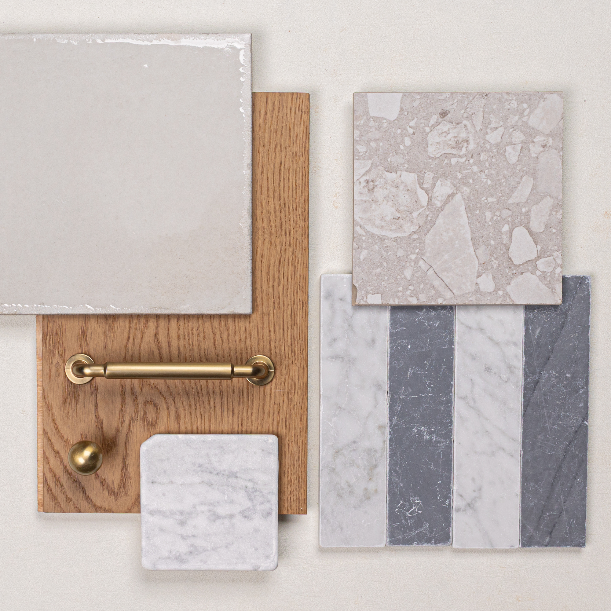Square ceramic and porcelain tile samples with oak wood and brass hardware pieces arranged on a neutral surface
