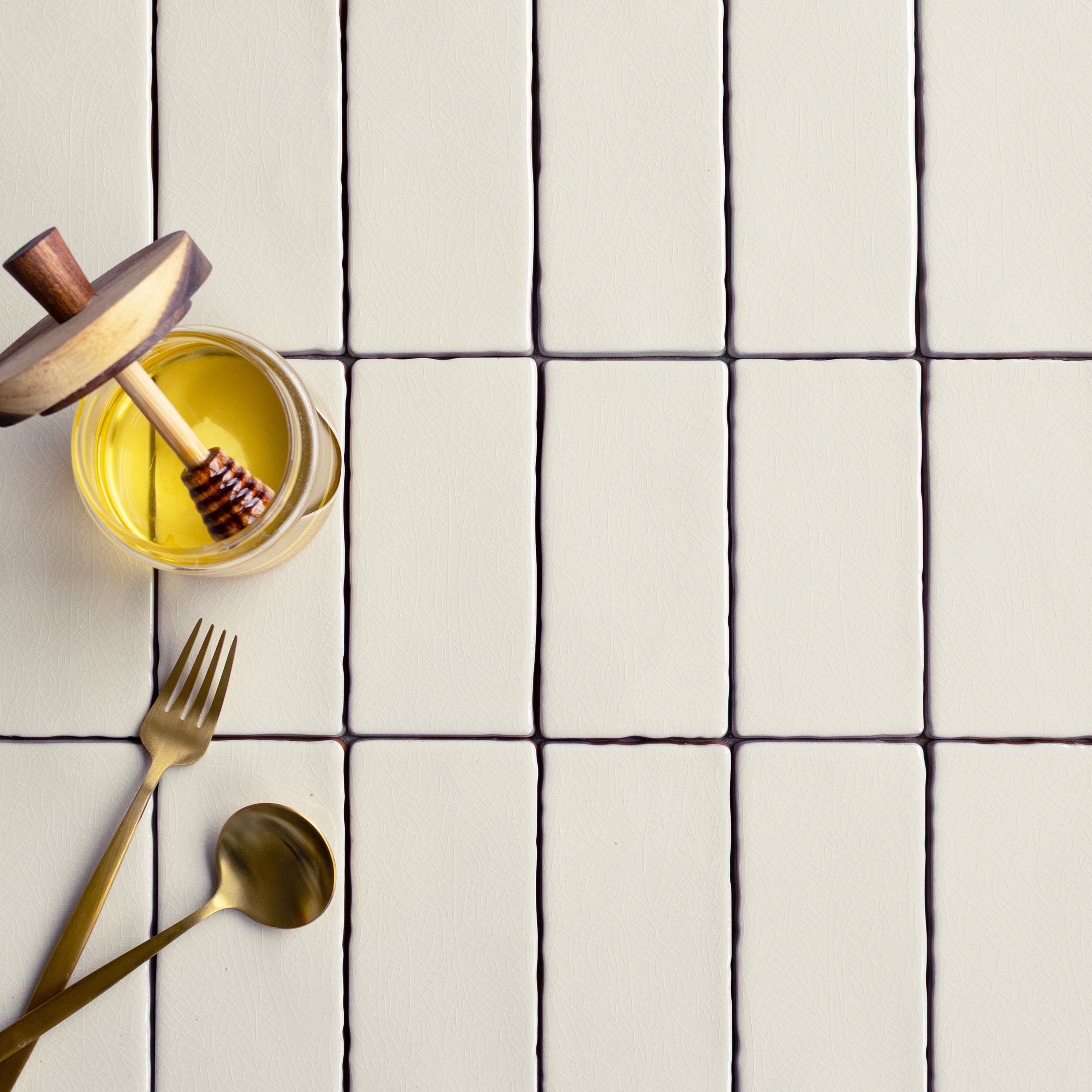 Matte off-white ceramic tiles with slightly uneven edges arranged in a grid beside gold-colored spoon and fork with honey jar