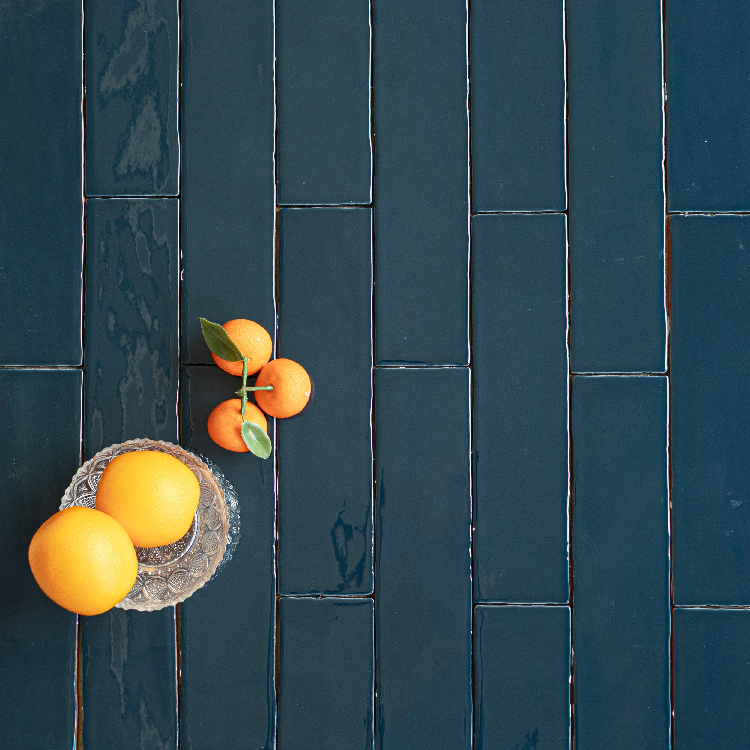 Glossy deep teal ceramic tiles with irregular edges arranged vertically alongside fresh oranges and a glass dish