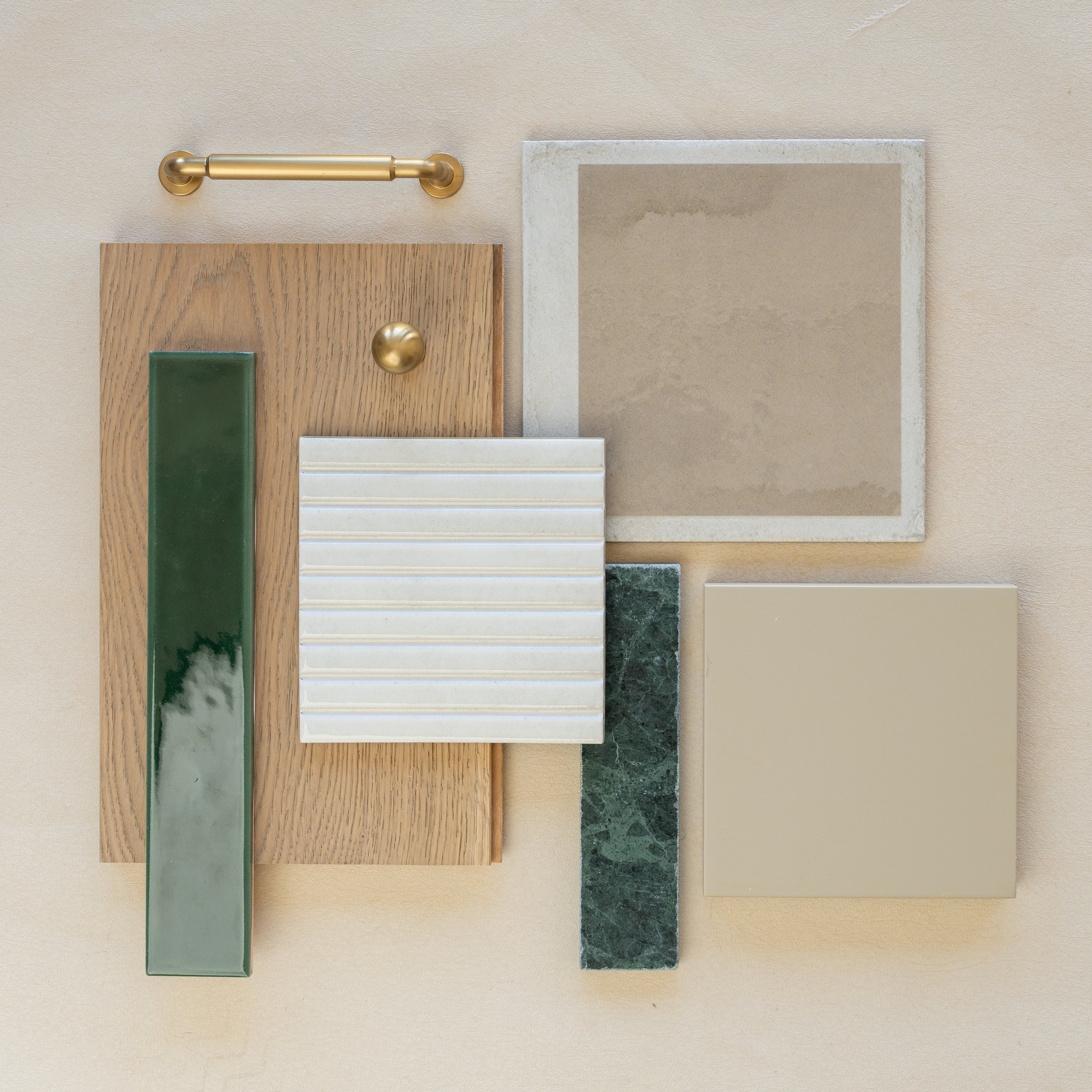 Flat lay of oak wood sample with brass hardware, beige square tiles, white ribbed ceramic tile, and green marble and ceramic tile pieces on neutral surface