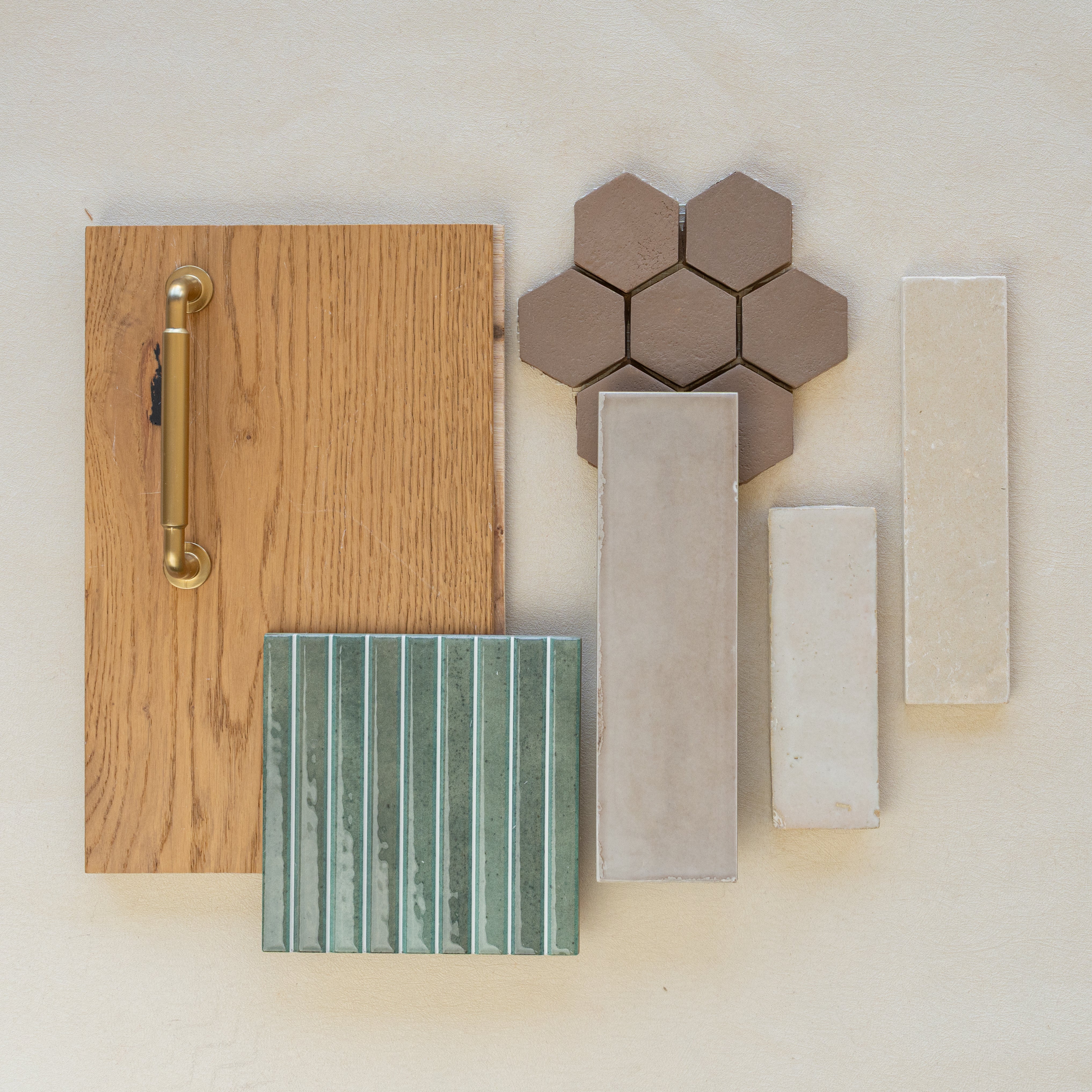 Flat lay of oak wood sample with brass handle, brown hexagonal tiles, green vertical ceramic tiles, and beige rectangular porcelain tiles on neutral background