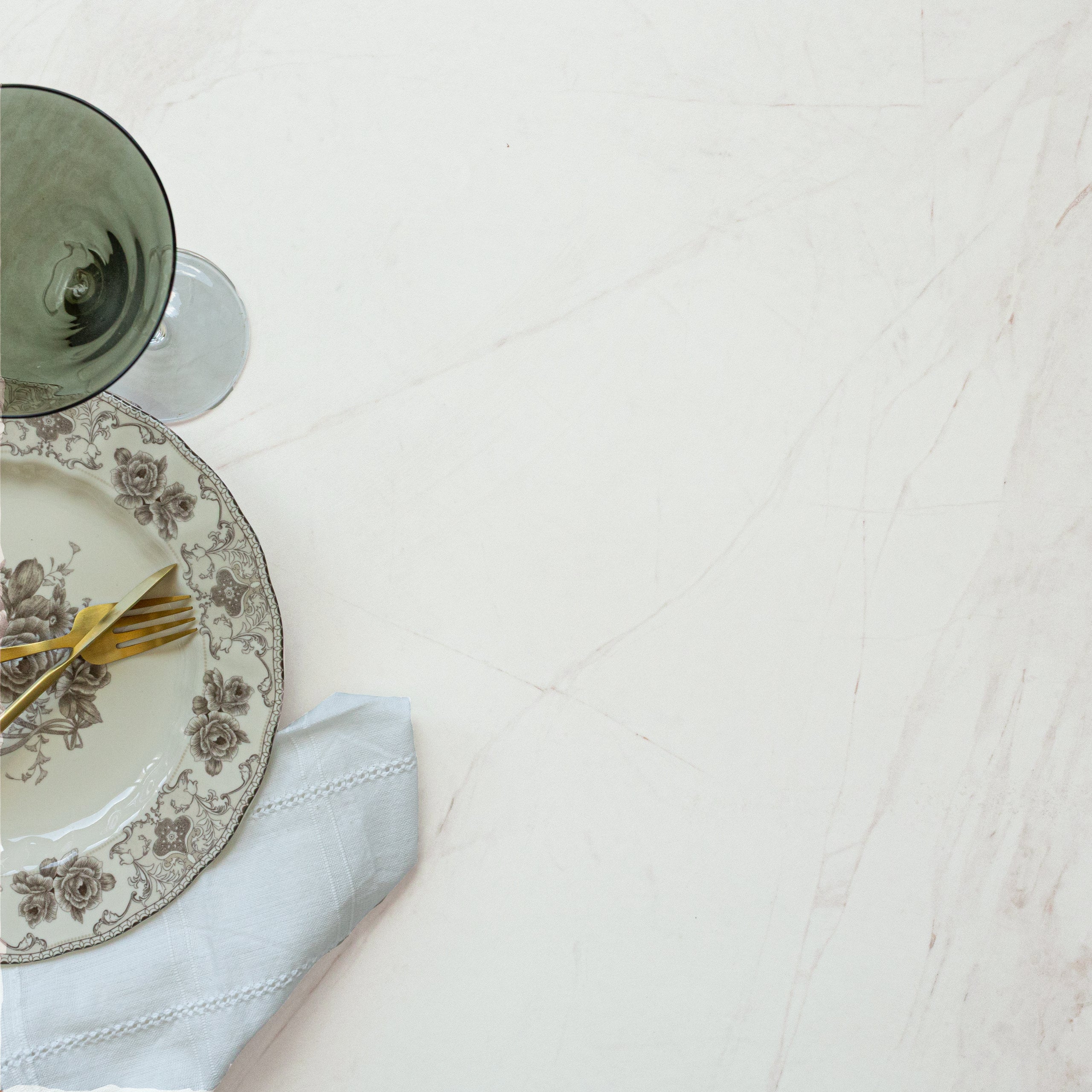 Vintage floral plate with gold flatware and green glass on smooth white porcelain tile with faint gold veining