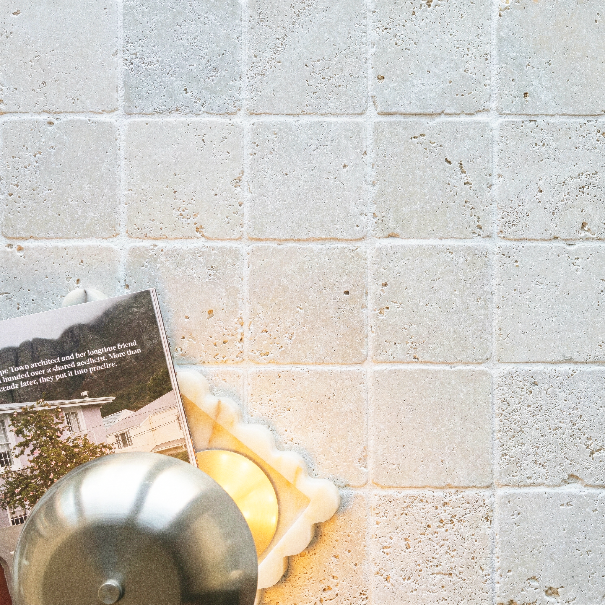 Light beige tumbled stone tiles with natural texture and subtle variations, accompanied by a metal teapot and magazine corner.
