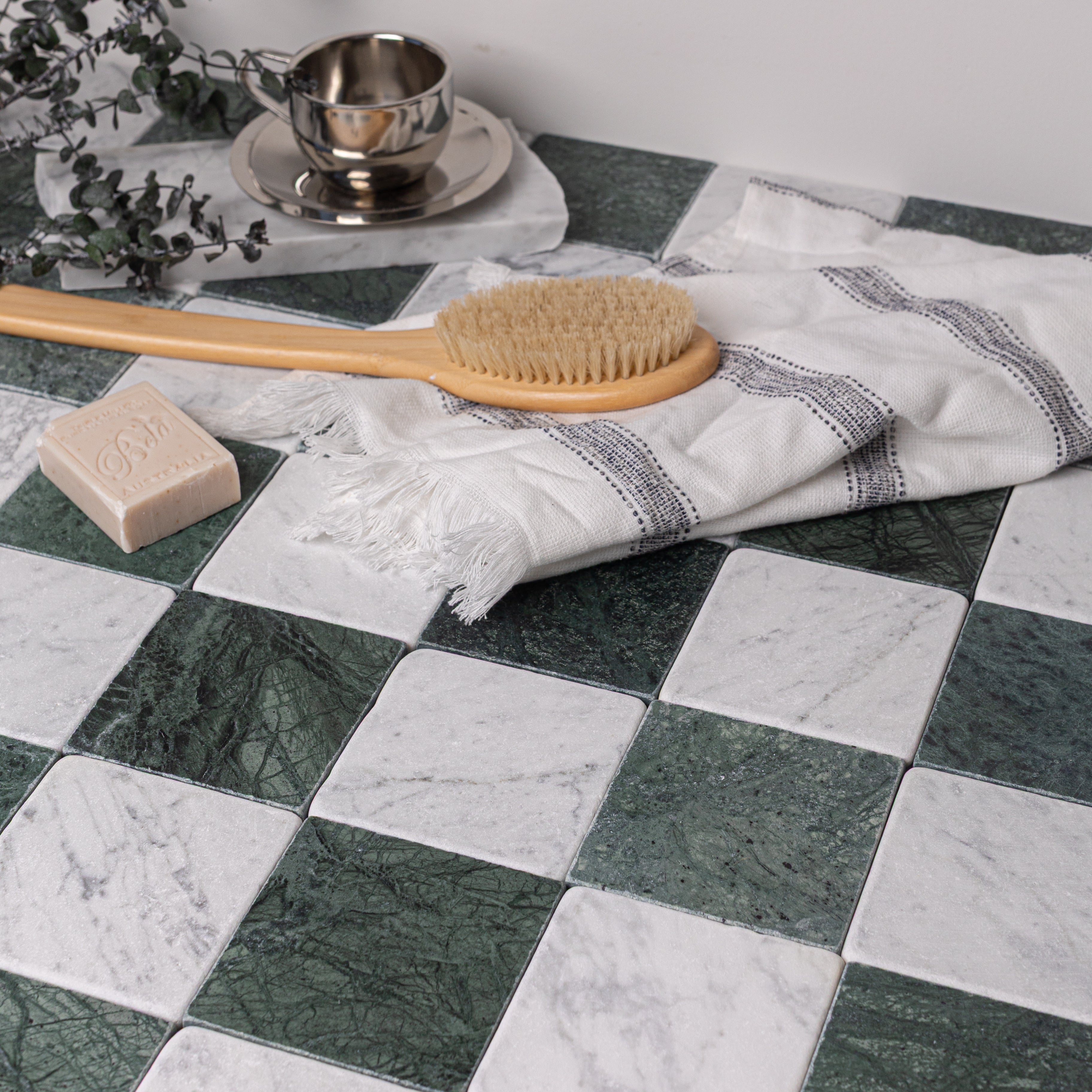Checkerboard arrangement of dark green and white tumbled marble tiles with linen cloth, brush, and soap bar on top