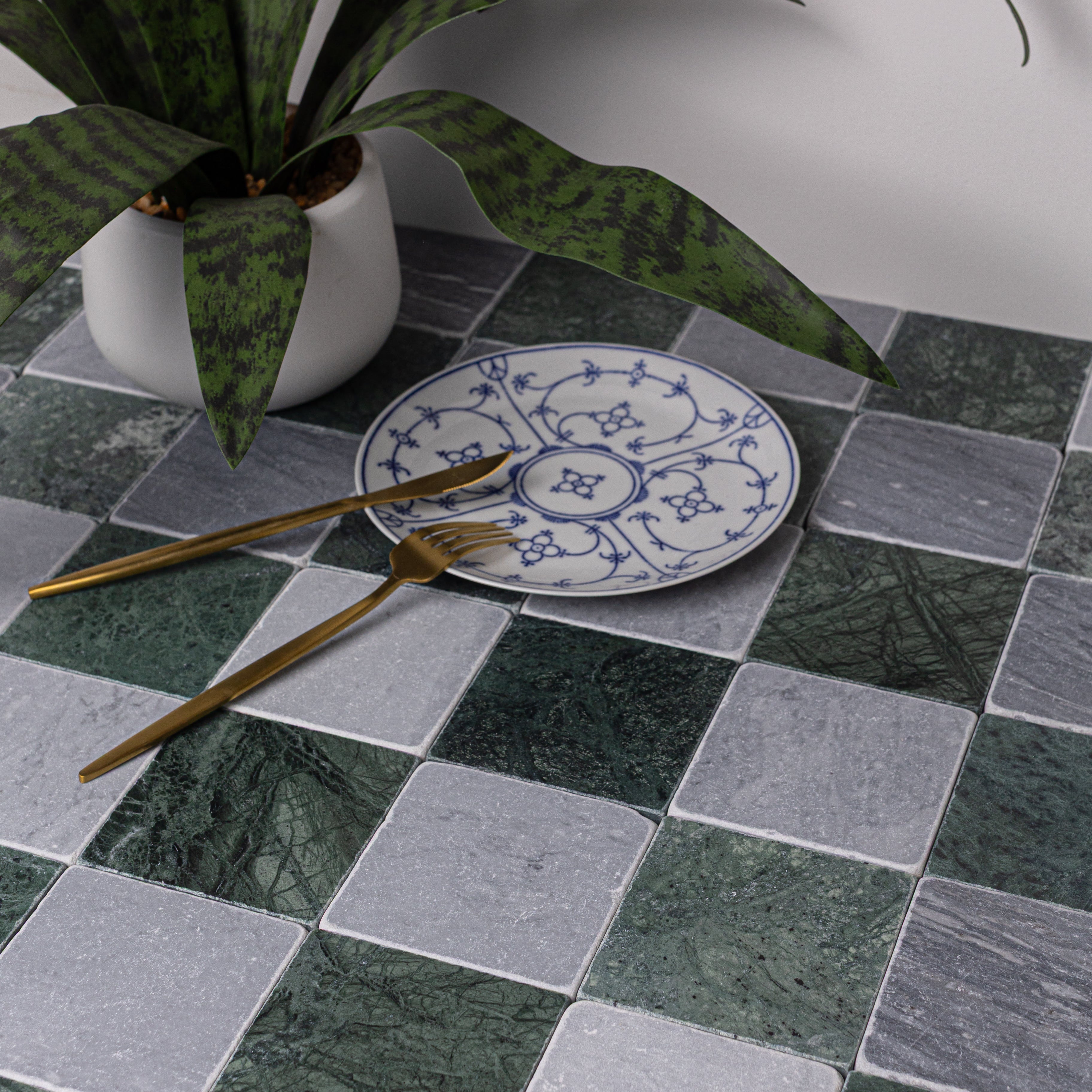Close-up of green and gray marble tiles with blue floral plate, gold fork and knife, and green spotted plant leaves above