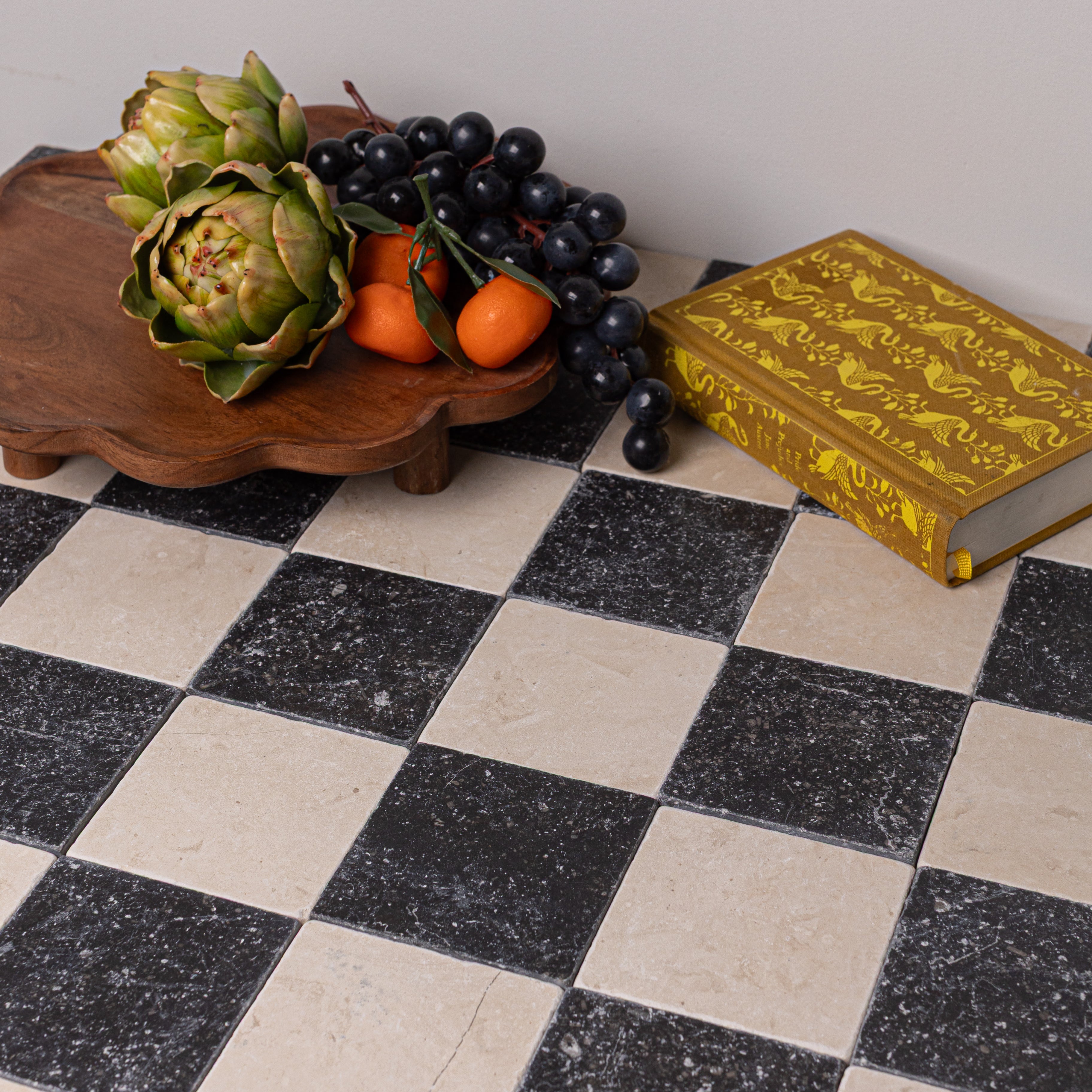 Checkerboard pattern of tumbled black and beige stone tiles with wooden tray, artichokes, grapes, and decorative book nearby