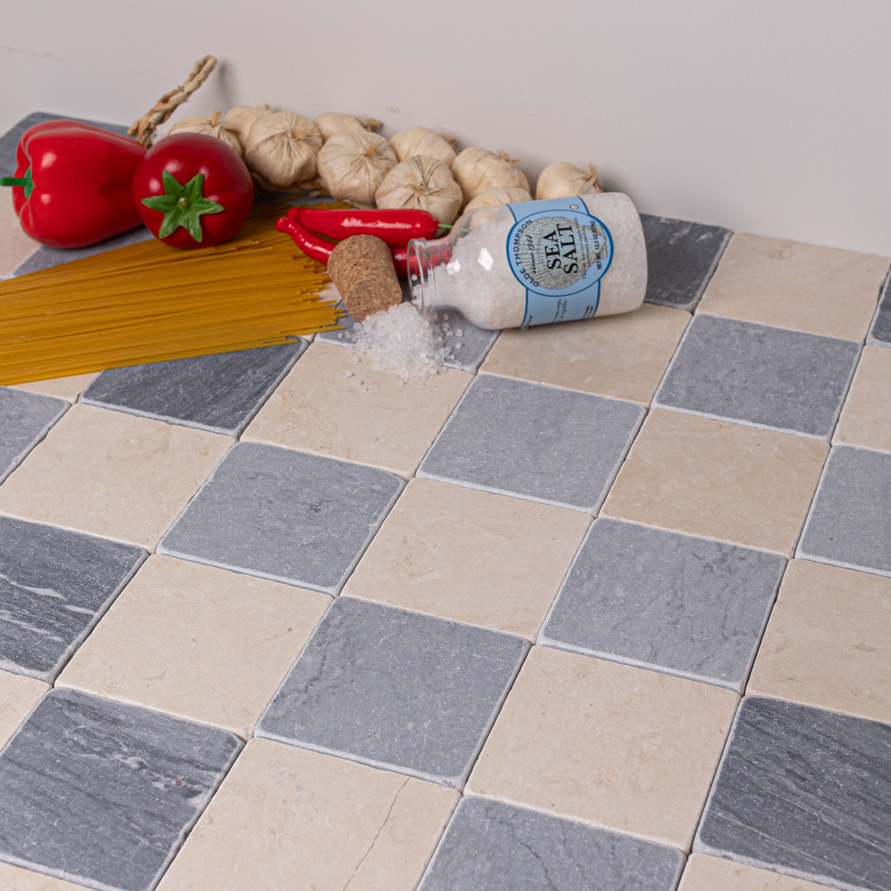 Checkerboard arrangement of beige and gray tumbled stone tiles with garlic, red pepper, spaghetti, and spilled sea salt jar