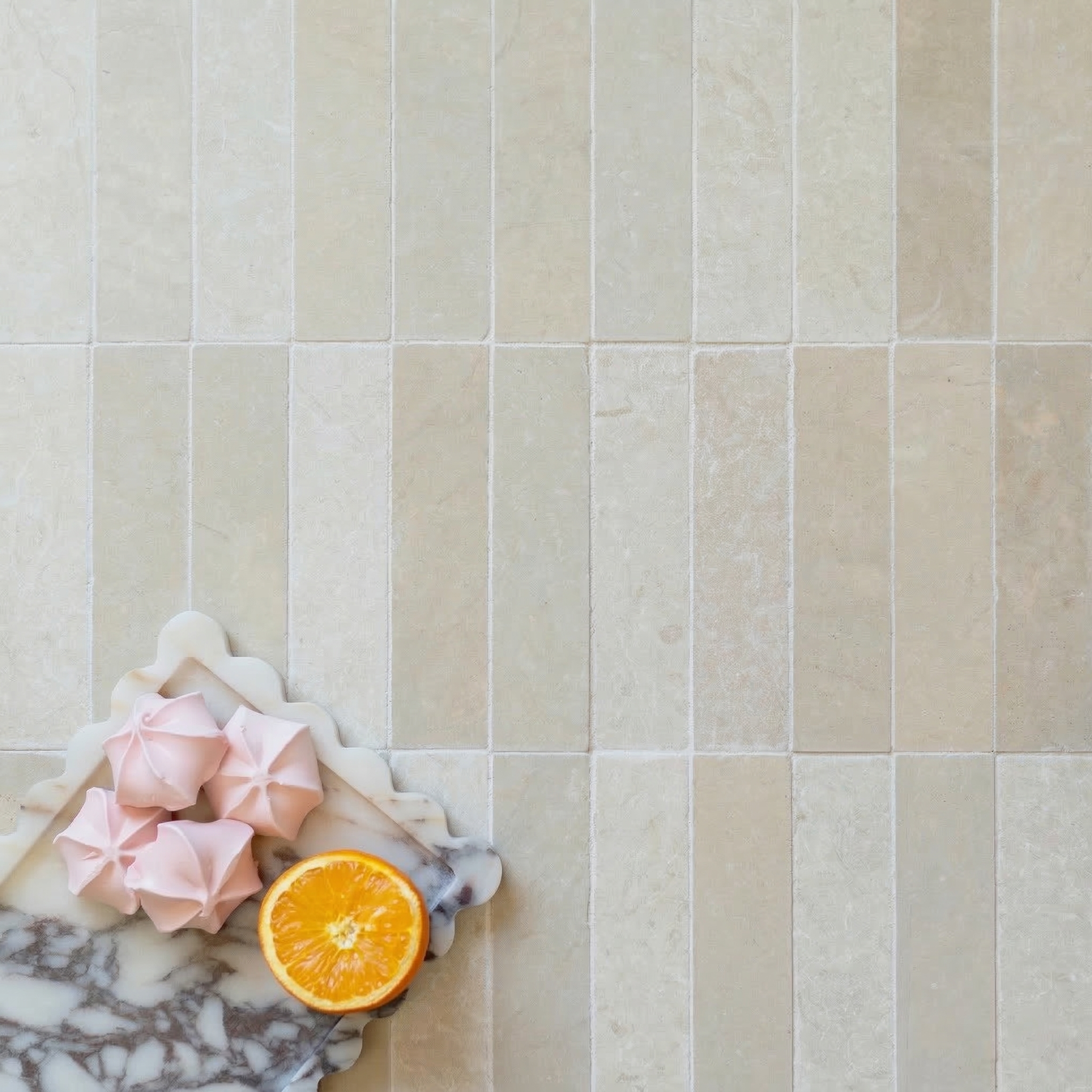 Soft beige rectangular stone tiles with natural texture arranged in vertical grid beneath marble tray with pink meringues and orange half