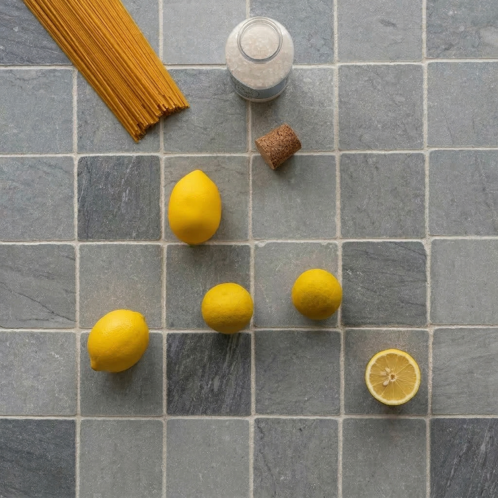 Natural gray stone tiles arranged in a grid with lemons, uncooked spaghetti, a cork, and a glass jar on top