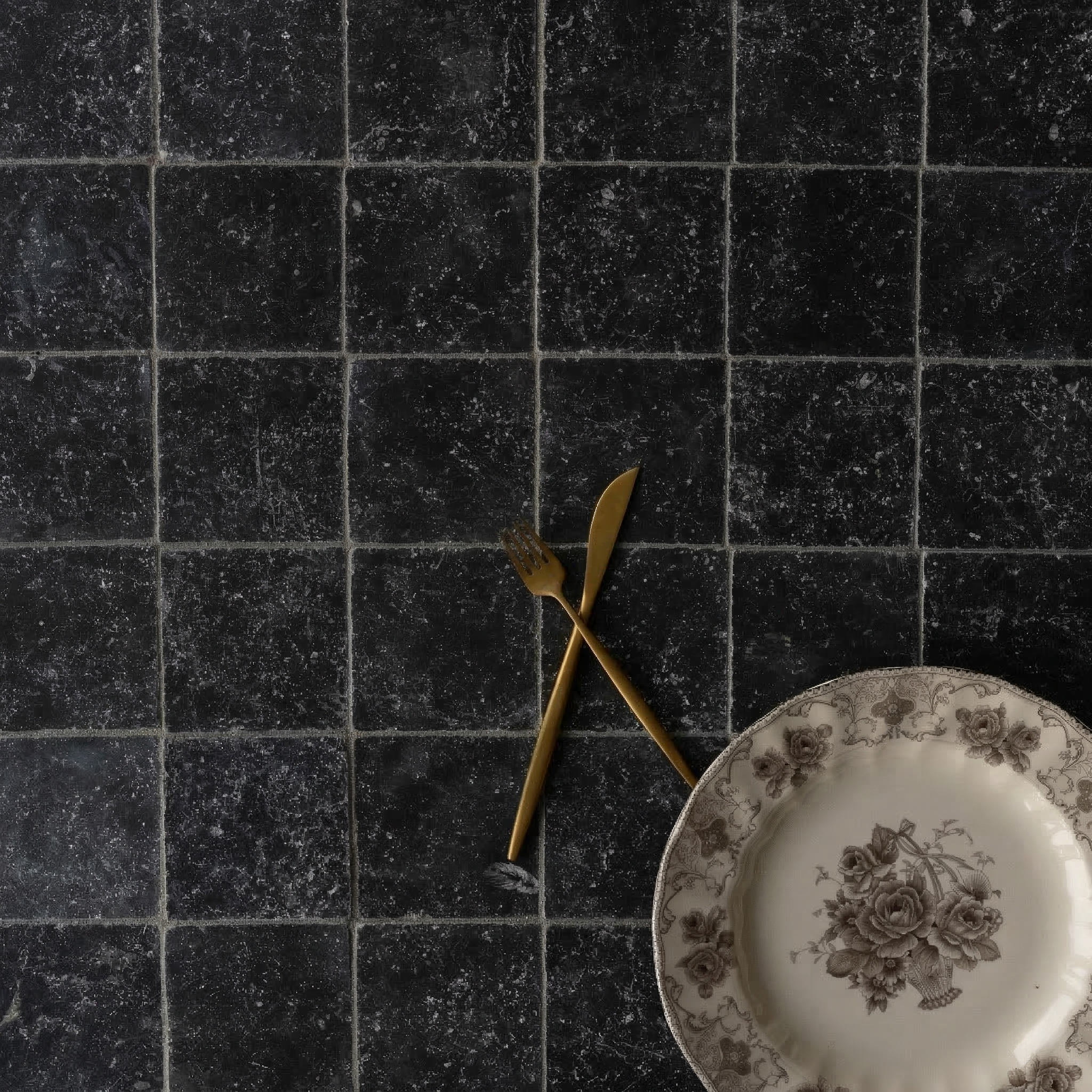 Dark square tumbled stone tiles with natural texture beneath a vintage floral plate and gold-toned fork and knife crossed.