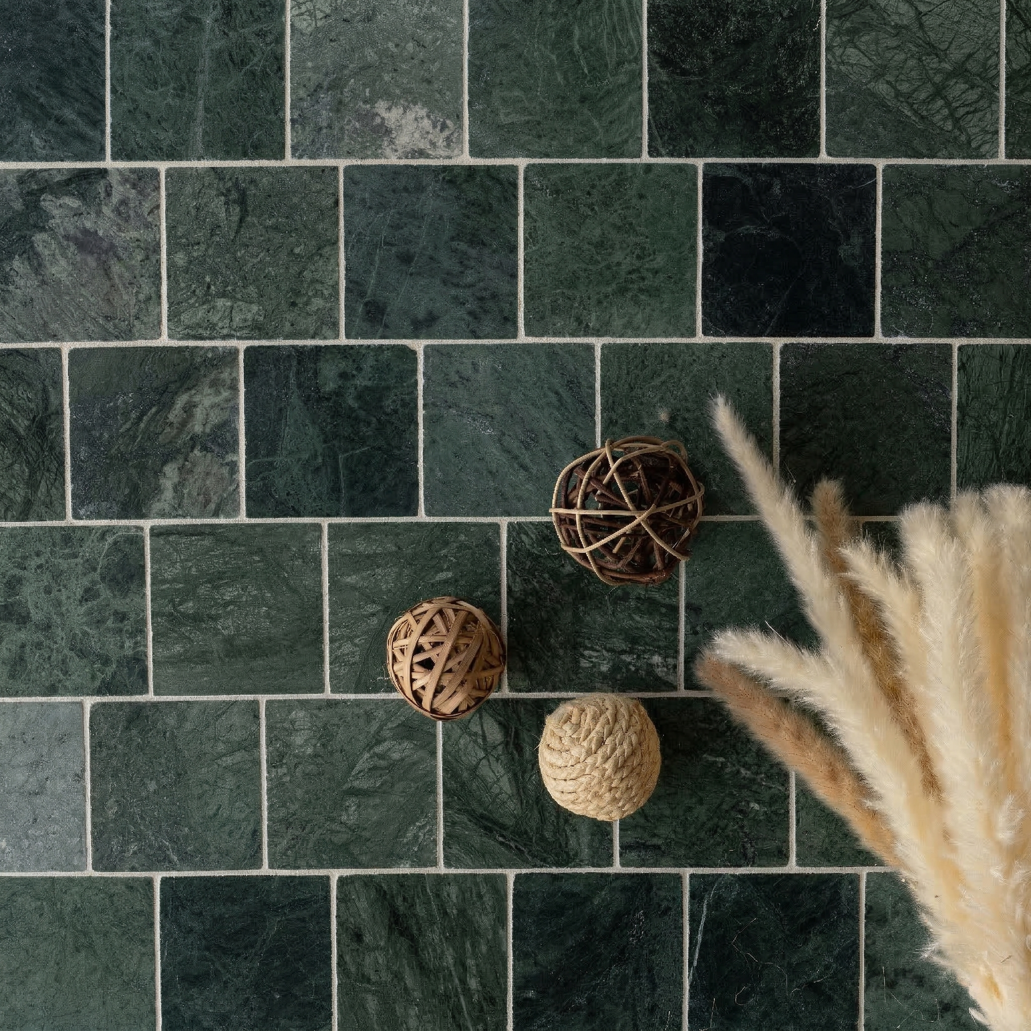 Square tumbled stone tiles in deep green shades with natural textures, paired with decorative wicker balls and pampas grass.