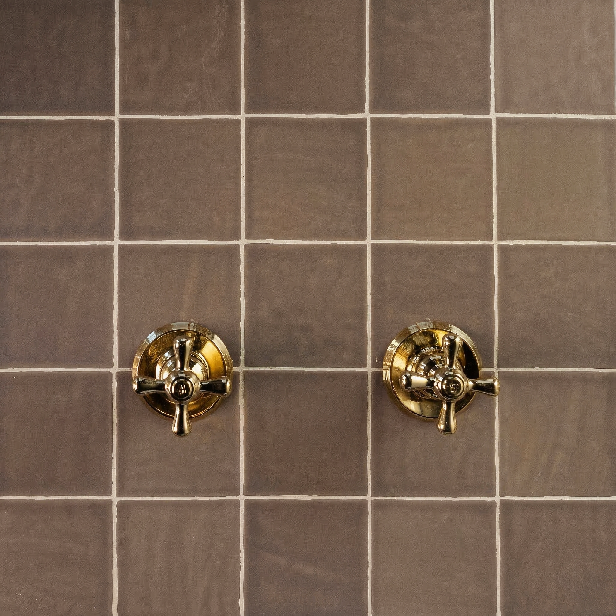 Glossy umber ceramic tiles with subtle texture and two polished brass shower valve handles on a smooth surface