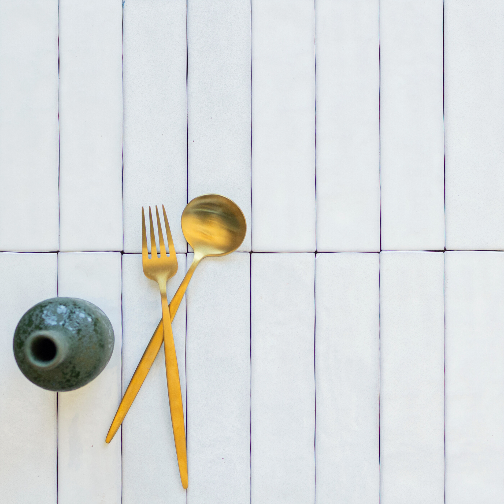 Matte white narrow rectangular ceramic tiles arranged vertically with a green ceramic vase and gold spoon and fork resting on top