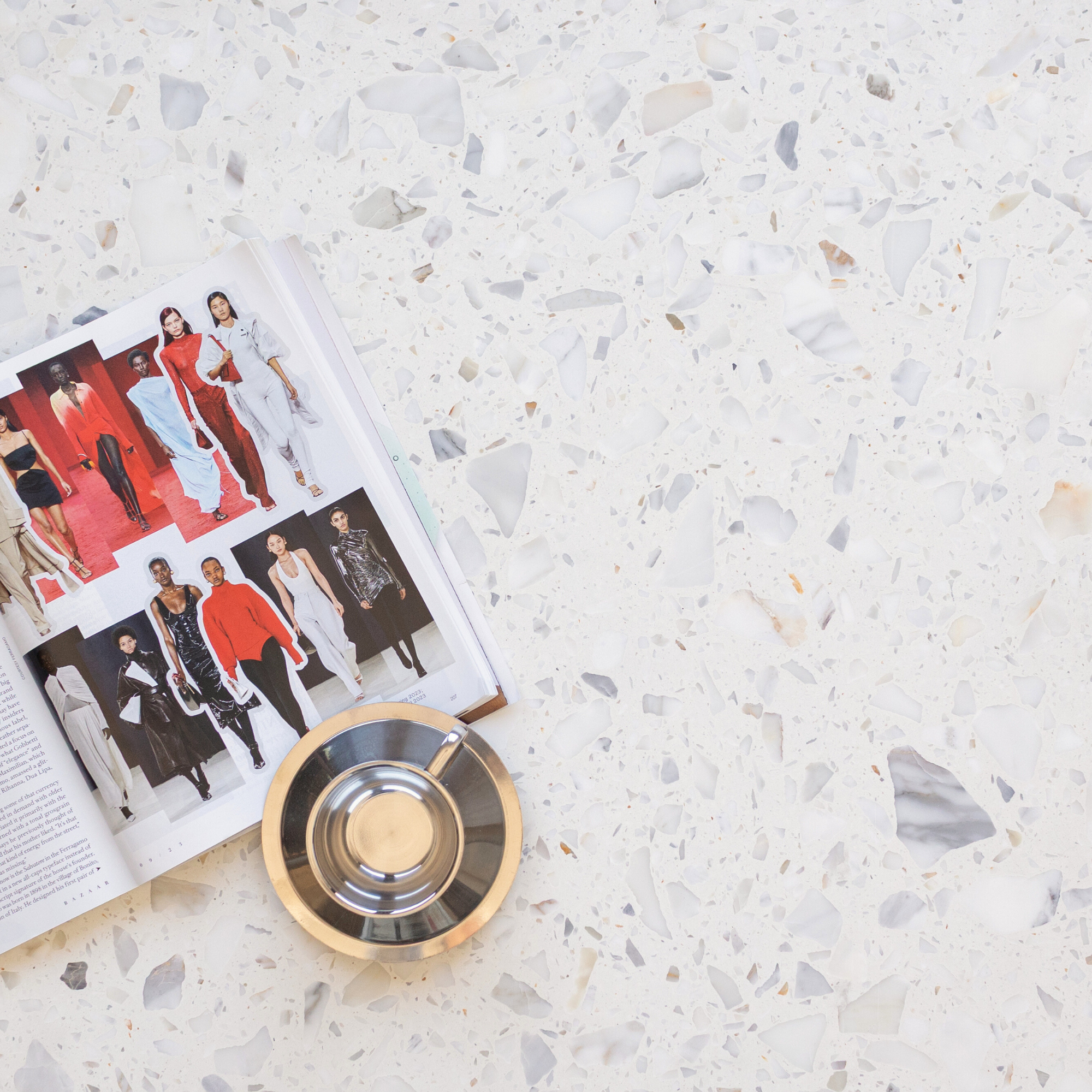 Open fashion magazine with runway images and a metallic ashtray resting on a white terrazzo surface with mixed stone chips