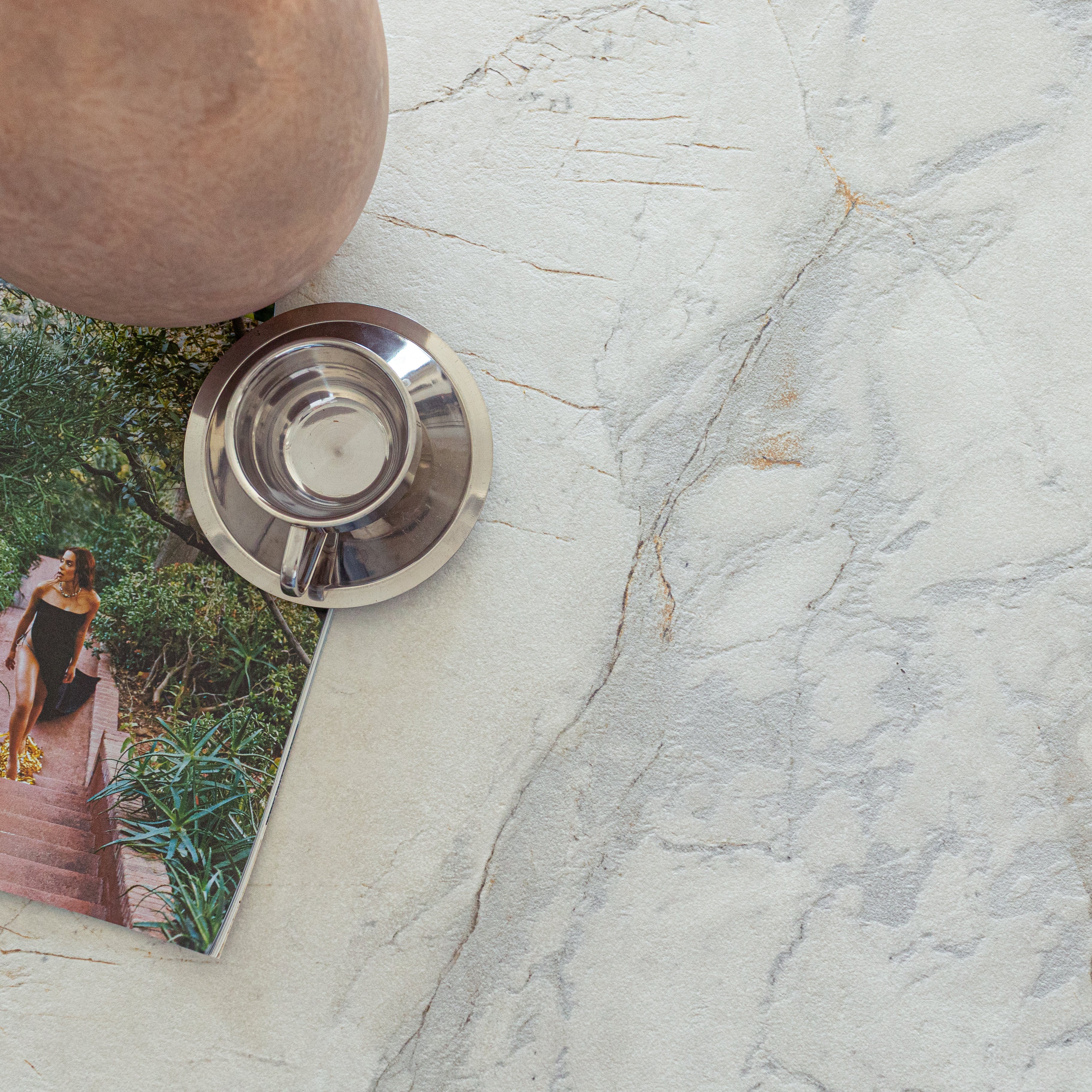 Light marble-effect porcelain tile surface with soft gray veining beside a metallic cup and an open magazine page