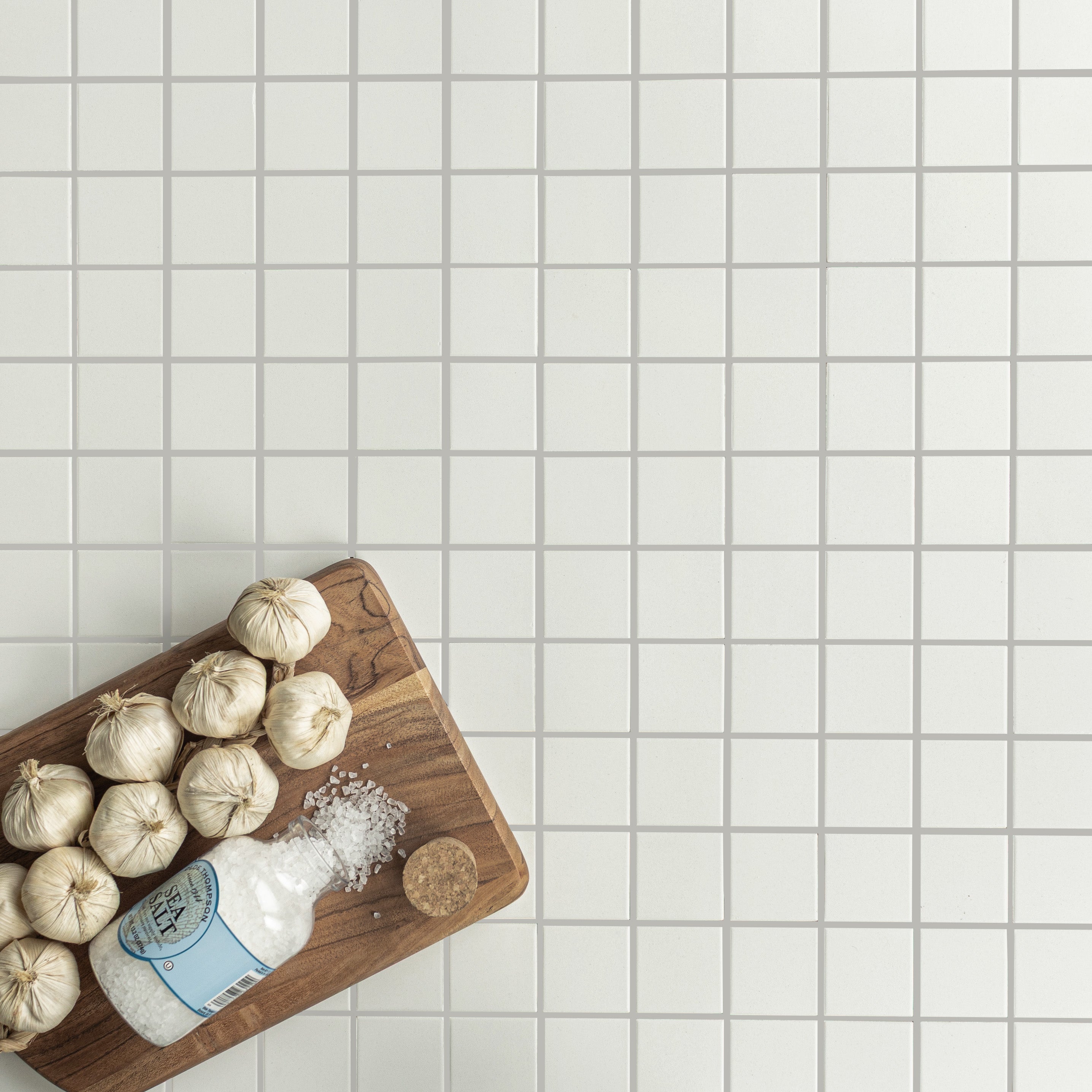 Overhead view of white mosaic ceramic tiles with wooden cutting board holding garlic bulbs and spilled sea salt jar