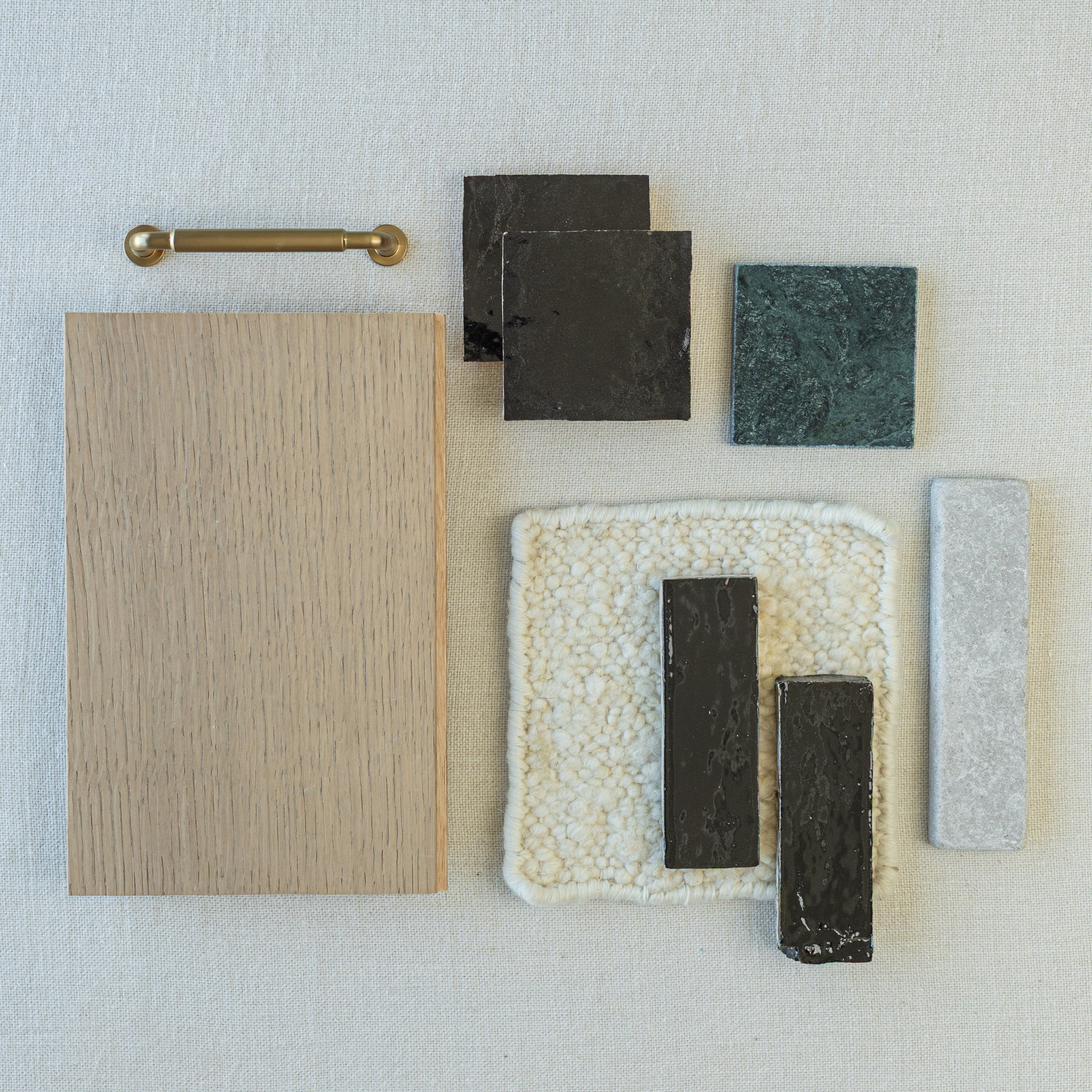 Flat lay of oak wood sample with brass handle, glossy black and textured green ceramic tiles, and soft cream fabric swatch