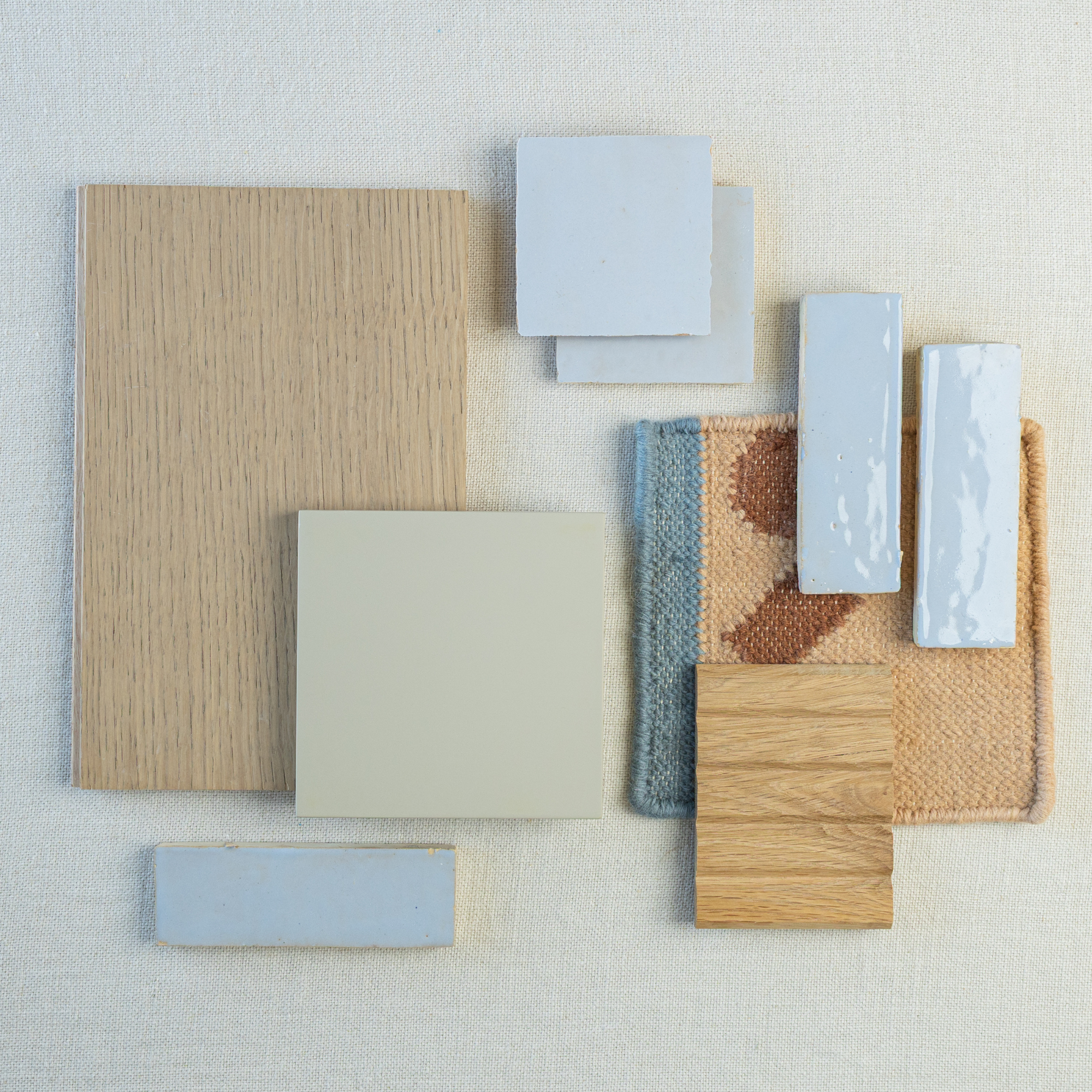 Flat lay of light oak wood samples, pale blue ceramic tiles, beige square tile, and textured fabric swatches on neutral background