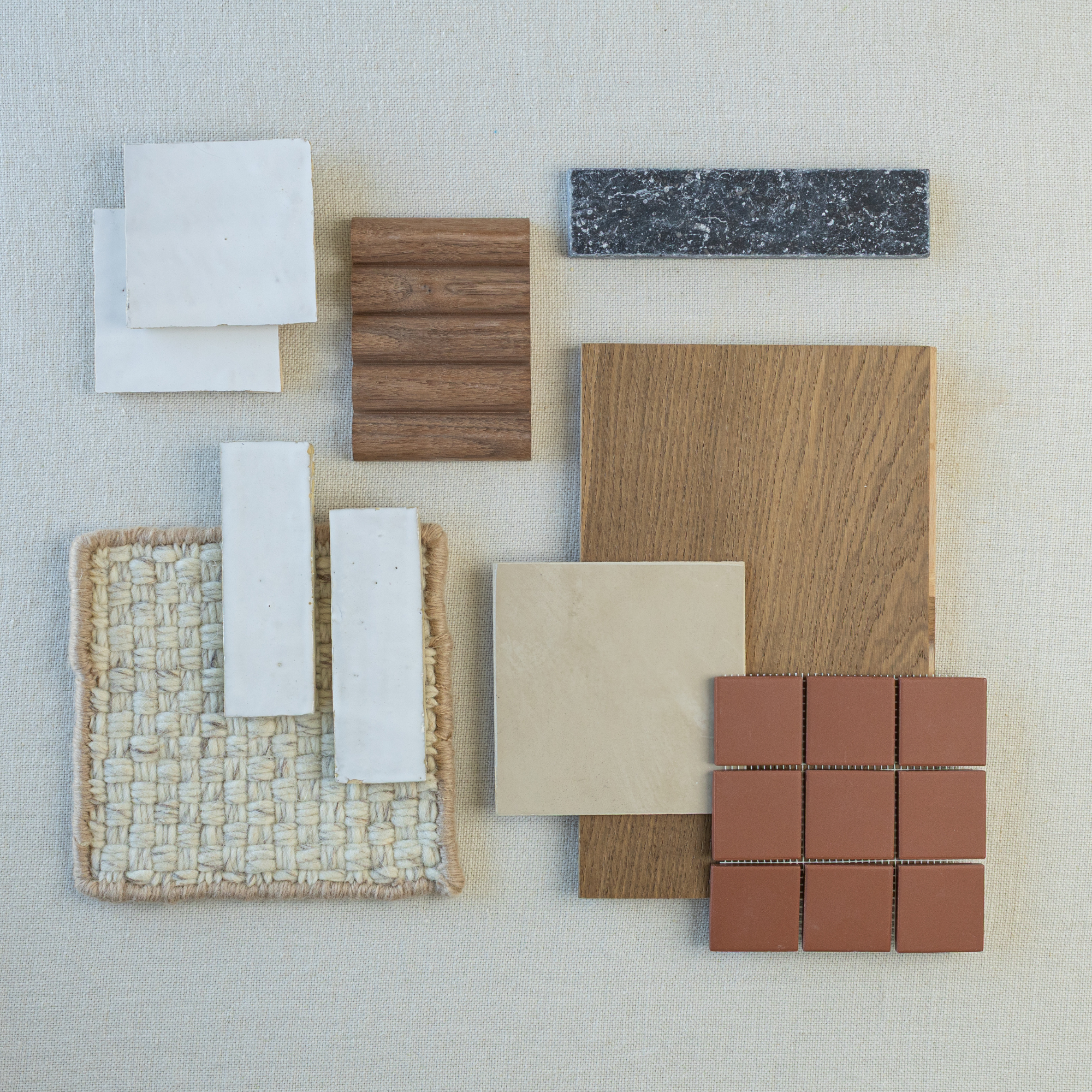 Flat lay of white ceramic tiles, warm wood samples, tan stone tile, woven fabric, dark stone slab, and terracotta square tiles on neutral background