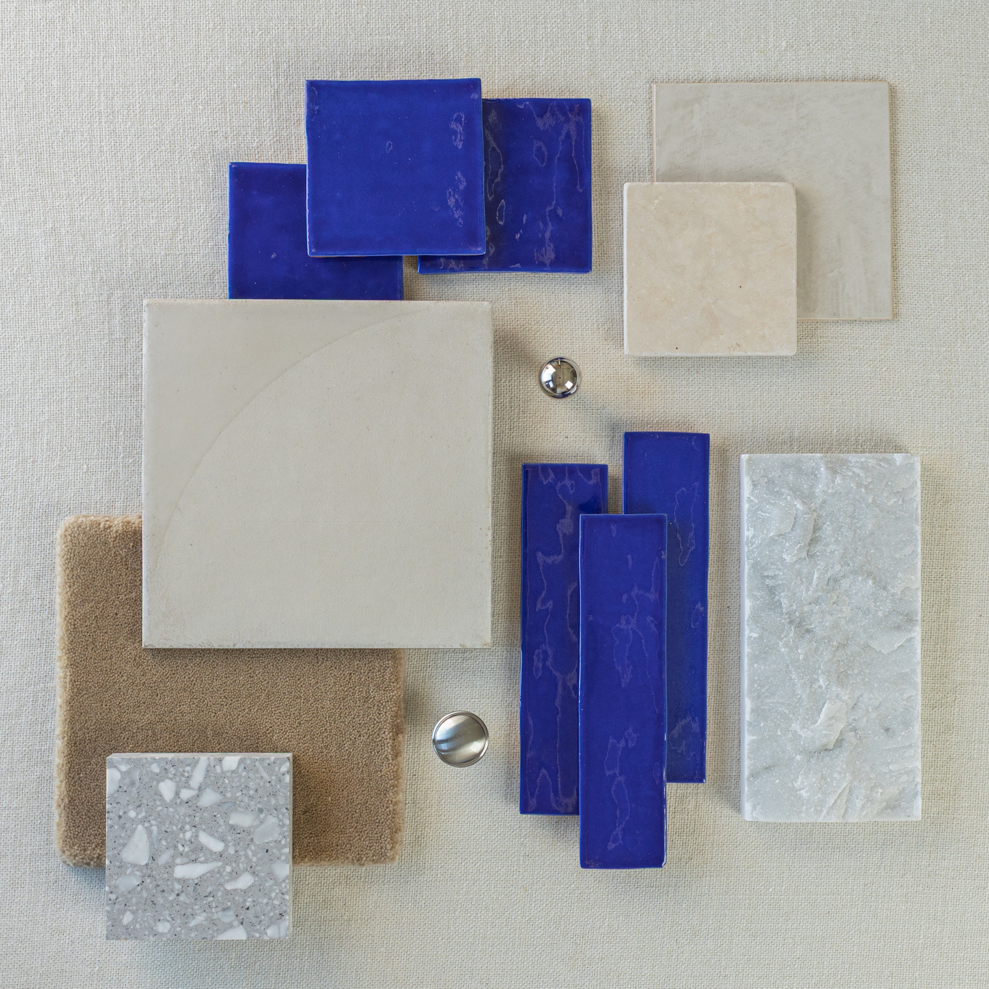 Flat lay of glossy cobalt blue ceramic tiles, beige stone tiles, textured fabric, and terrazzo sample on neutral fabric background