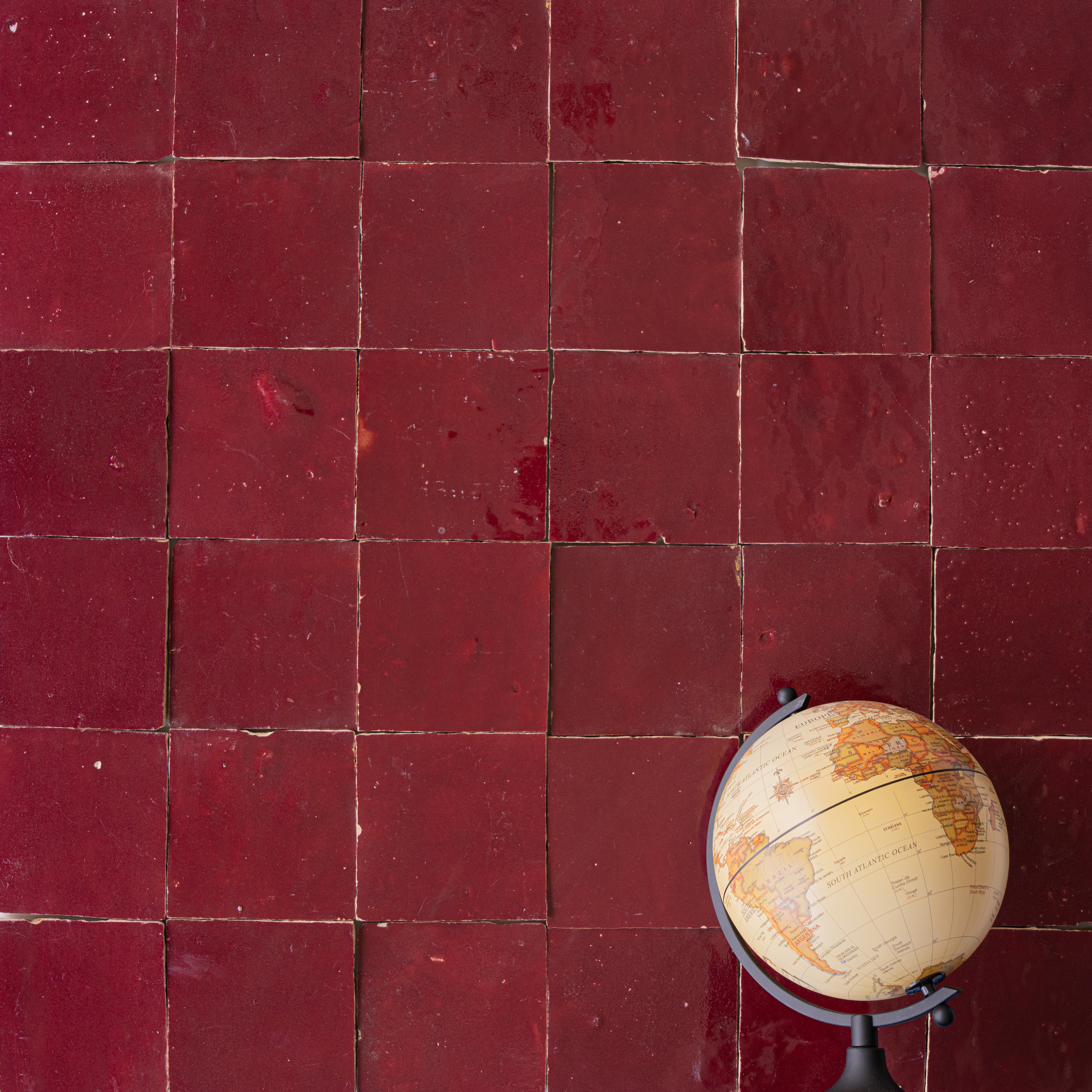 Deep red ceramic zellige tiles with a glossy finish arranged in a grid, accompanied by a small vintage globe on the corner