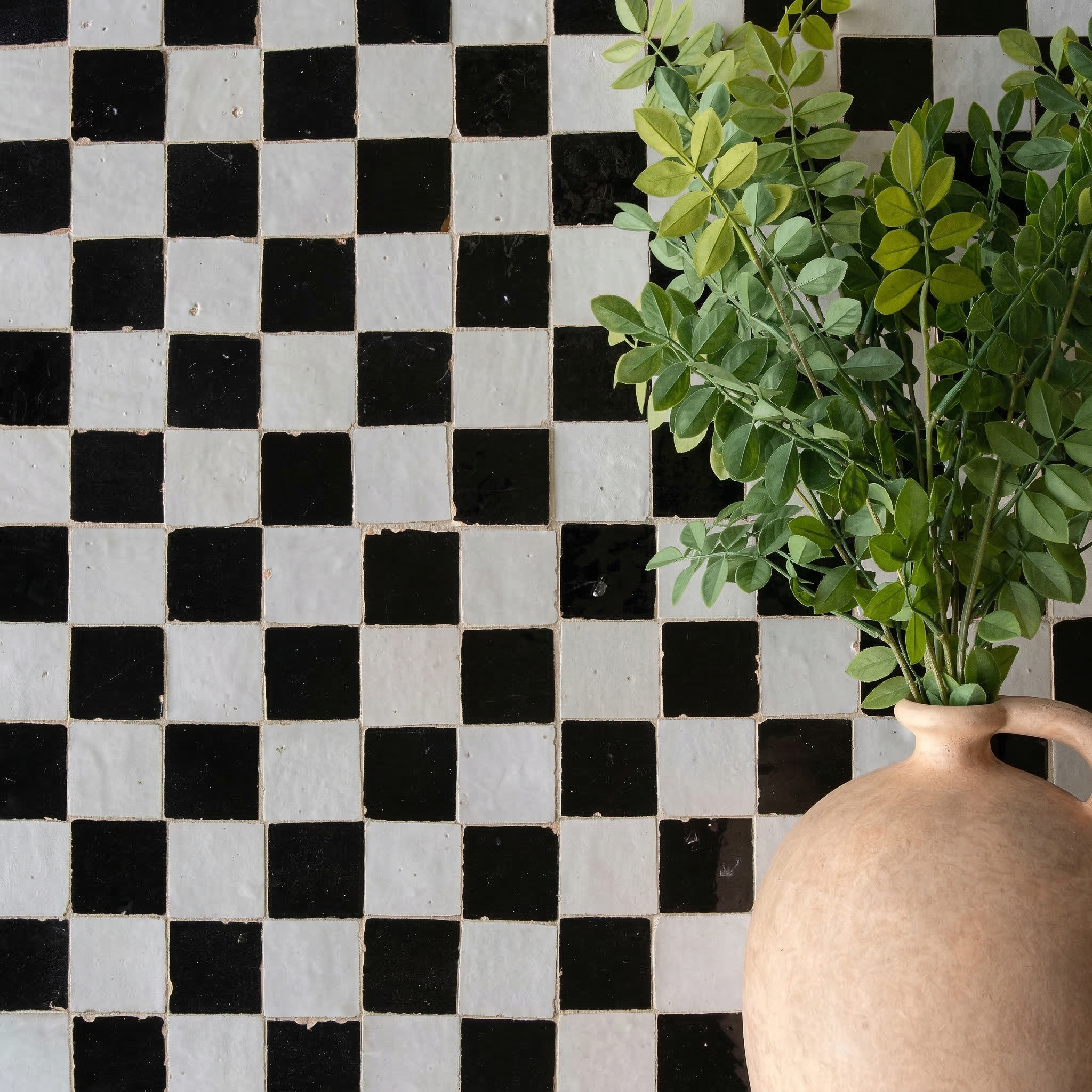 Checkerboard patterned small square black and white Zellige tiles with a rustic clay vase holding leafy green stems nearby