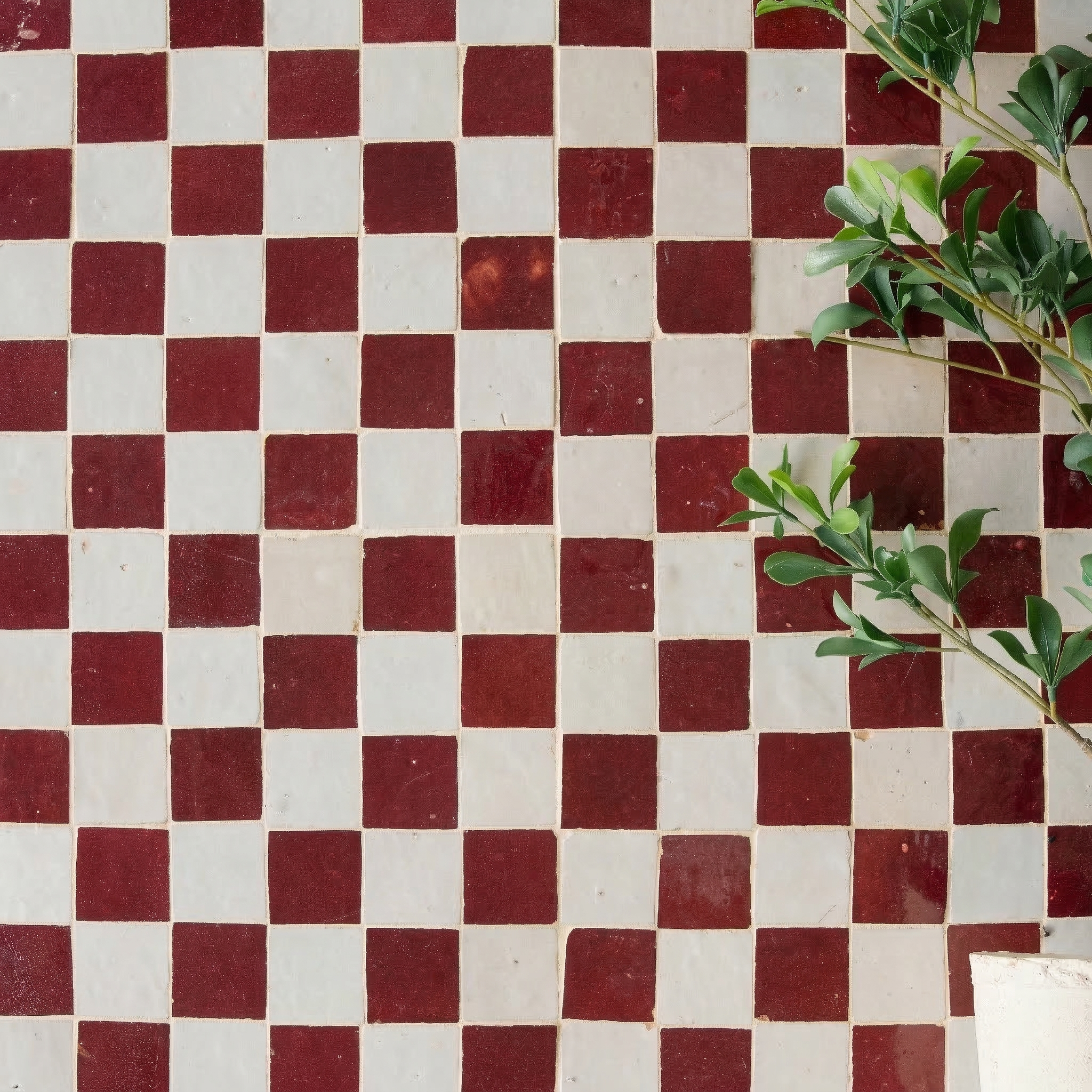 Handcrafted small square Zellige tiles in rich oxblood red and white arranged in a checkerboard pattern with greenery accents