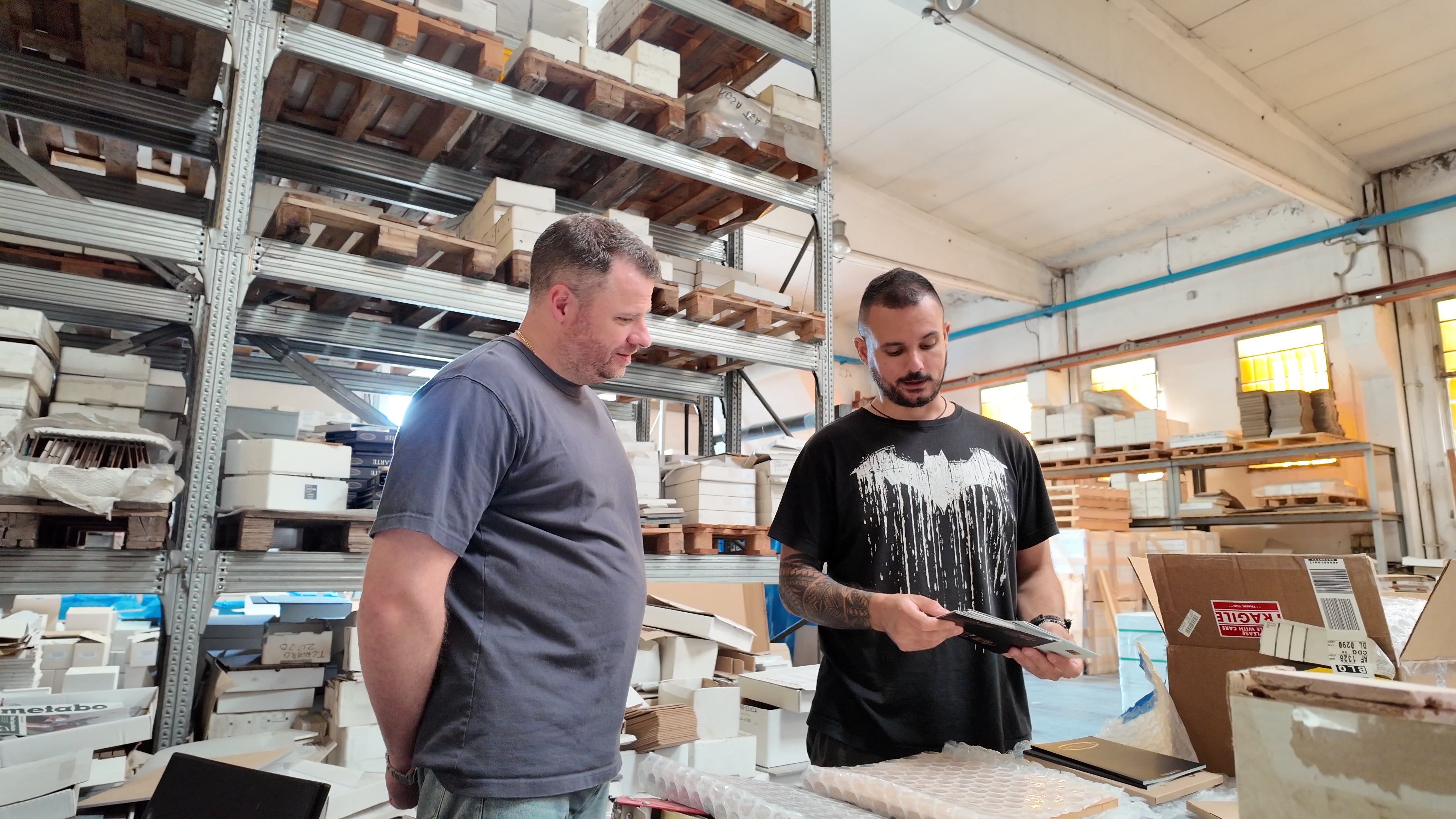 Two men examining ceramic tiles in a bright warehouse filled with stacked shelves and natural light