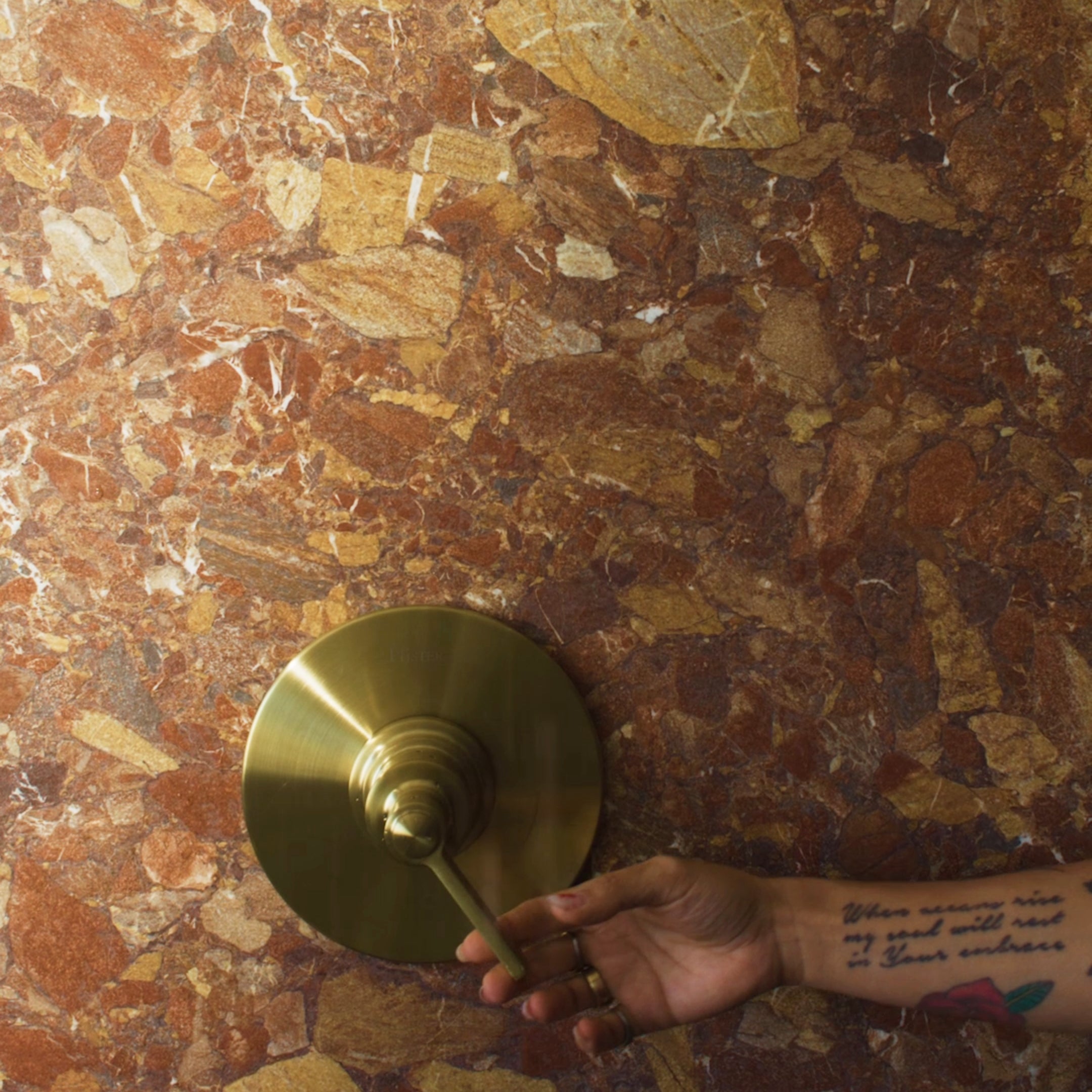 Hand adjusting antique brass faucet handle mounted on polished reddish-brown natural stone wall with irregular veining