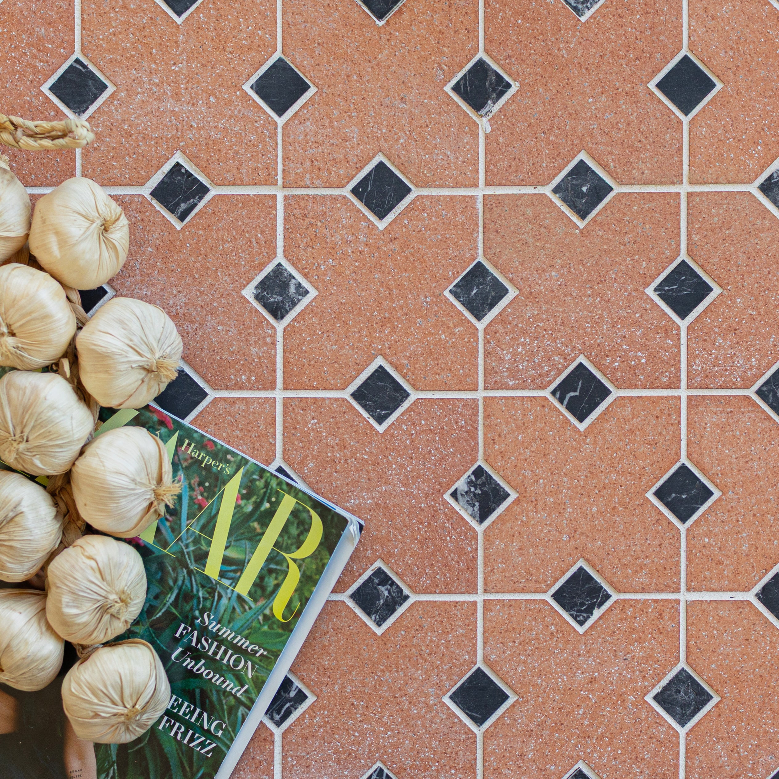 Terracotta ceramic tiles with diamond-shaped black accents arranged in a geometric pattern with garlic bulbs and a magazine nearby