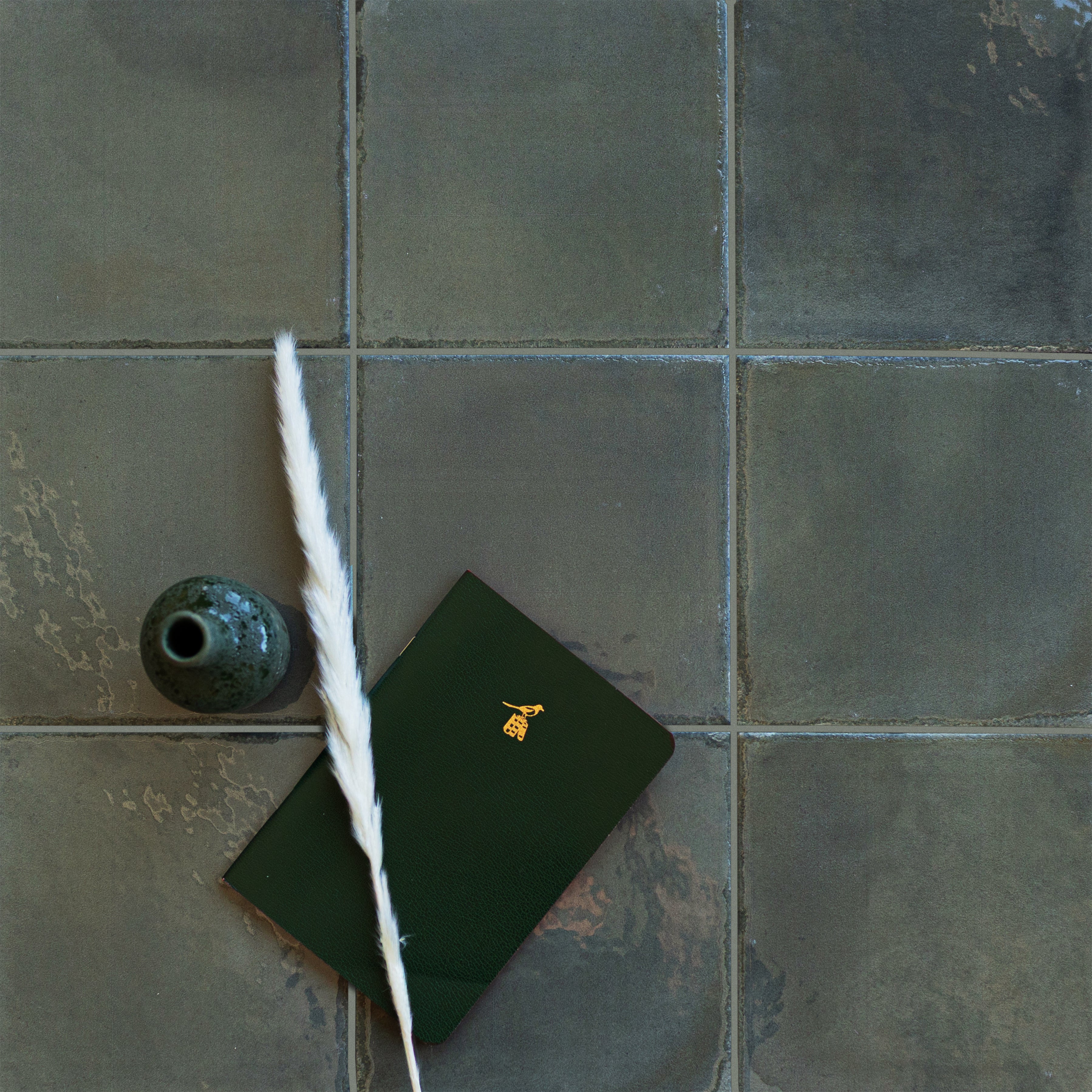Muted green ceramic tiles with a small dark vase, eucalyptus leaf, and green notebook on a textured surface
