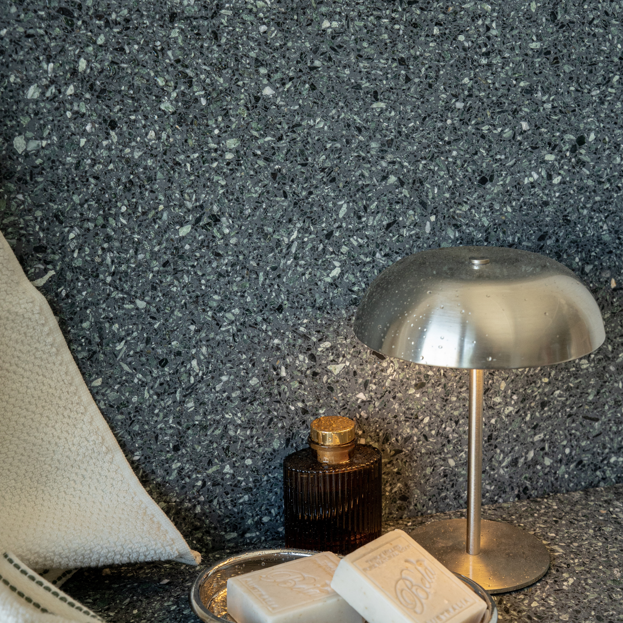 Dark terrazzo wall and counter with a metal dome lamp soap bars amber bottle and folded fabric