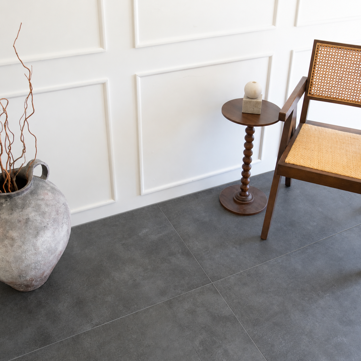 Large gray floor tiles in a styled room with a woven chair wooden side table and ceramic vase