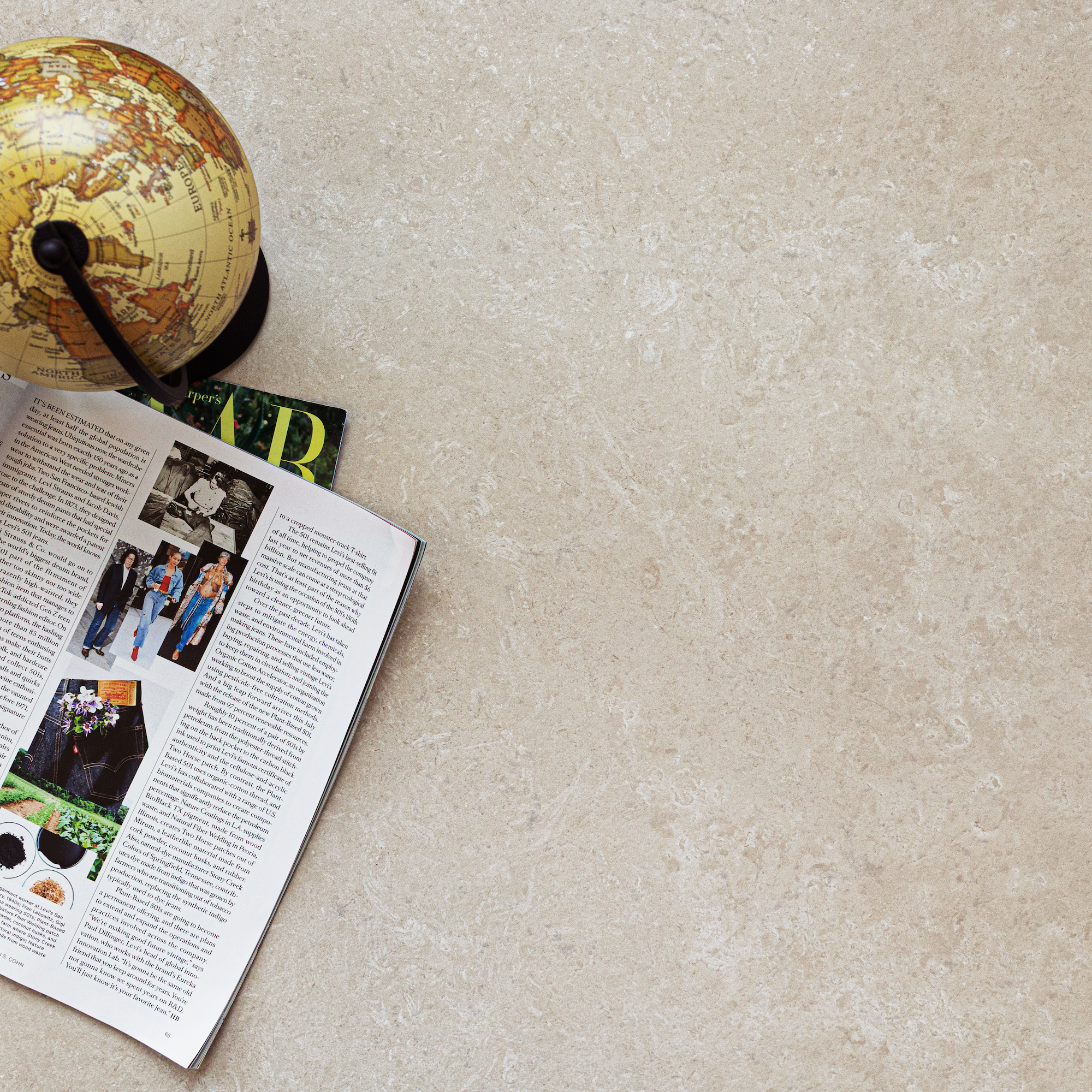 Cream-colored large format tile surface with natural stone texture beside an open magazine and vintage globe on neutral background